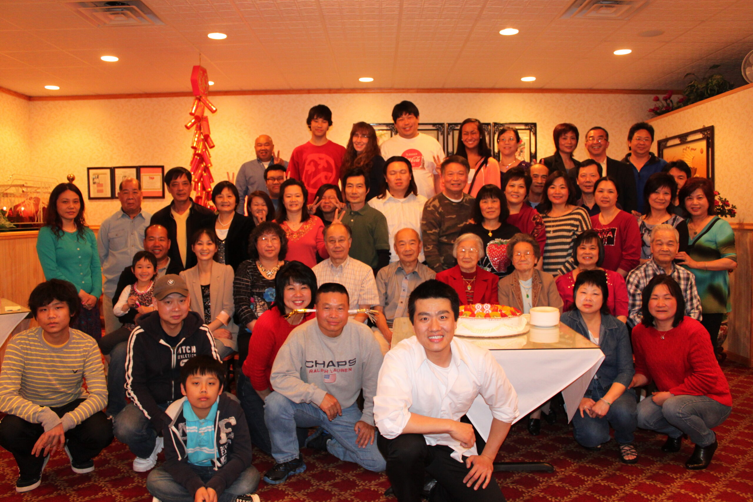 Large group of people posing for a photo at a birthday party around a cake on a table in the foreground.