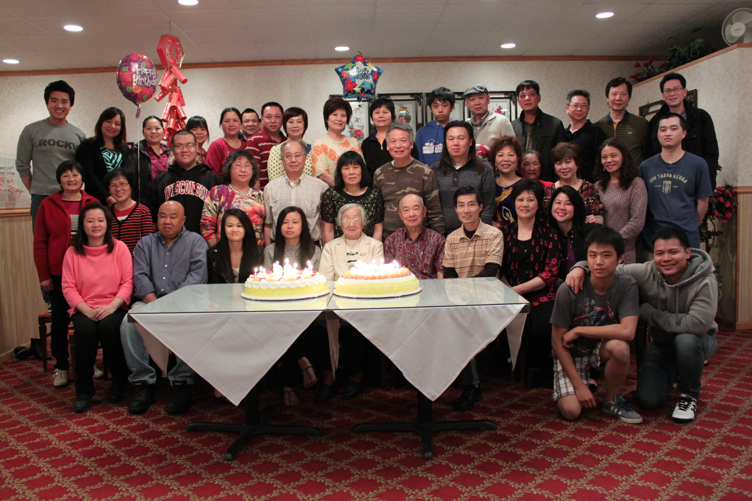 Large group of people gathered for a birthday celebration around two cakes on a table, with balloons overhead in a banquet room.