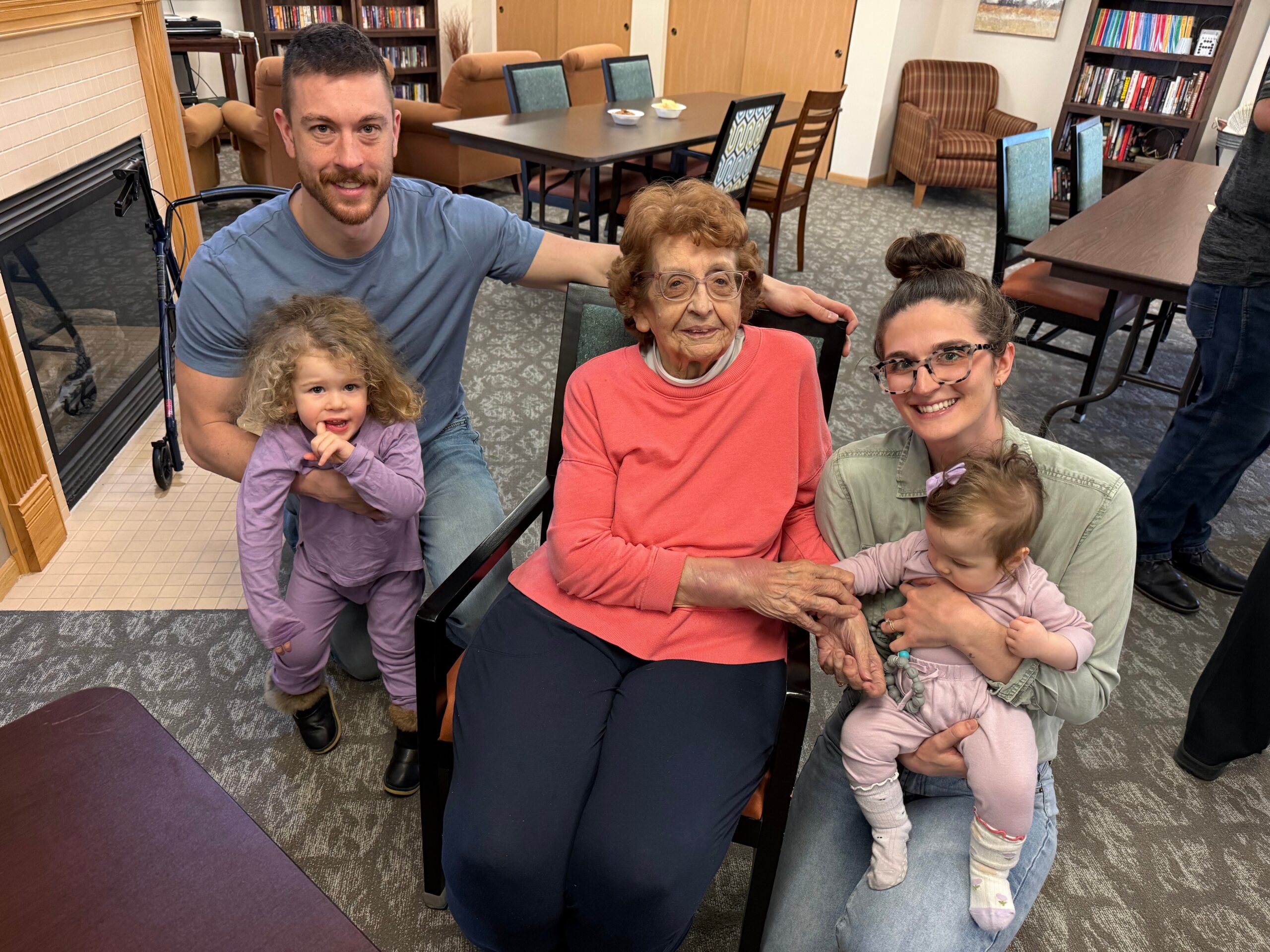 Family photo in a library lounge: elderly woman in a pink sweater is seated in a wheelchair, flanked by adults and young children, all smiling.
