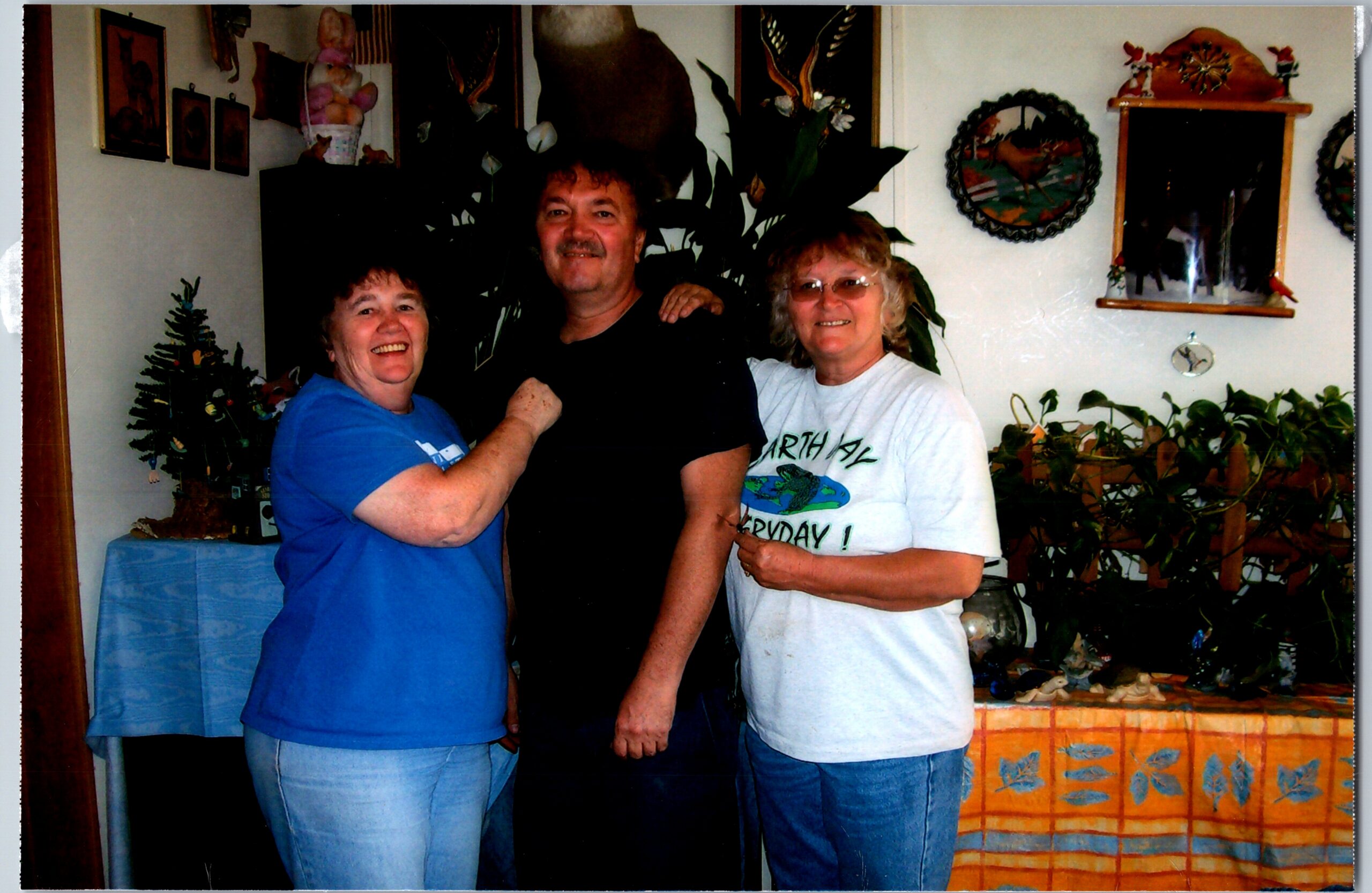 Three adults pose together indoors, smiling, with plants and wall decorations in a living room behind them.