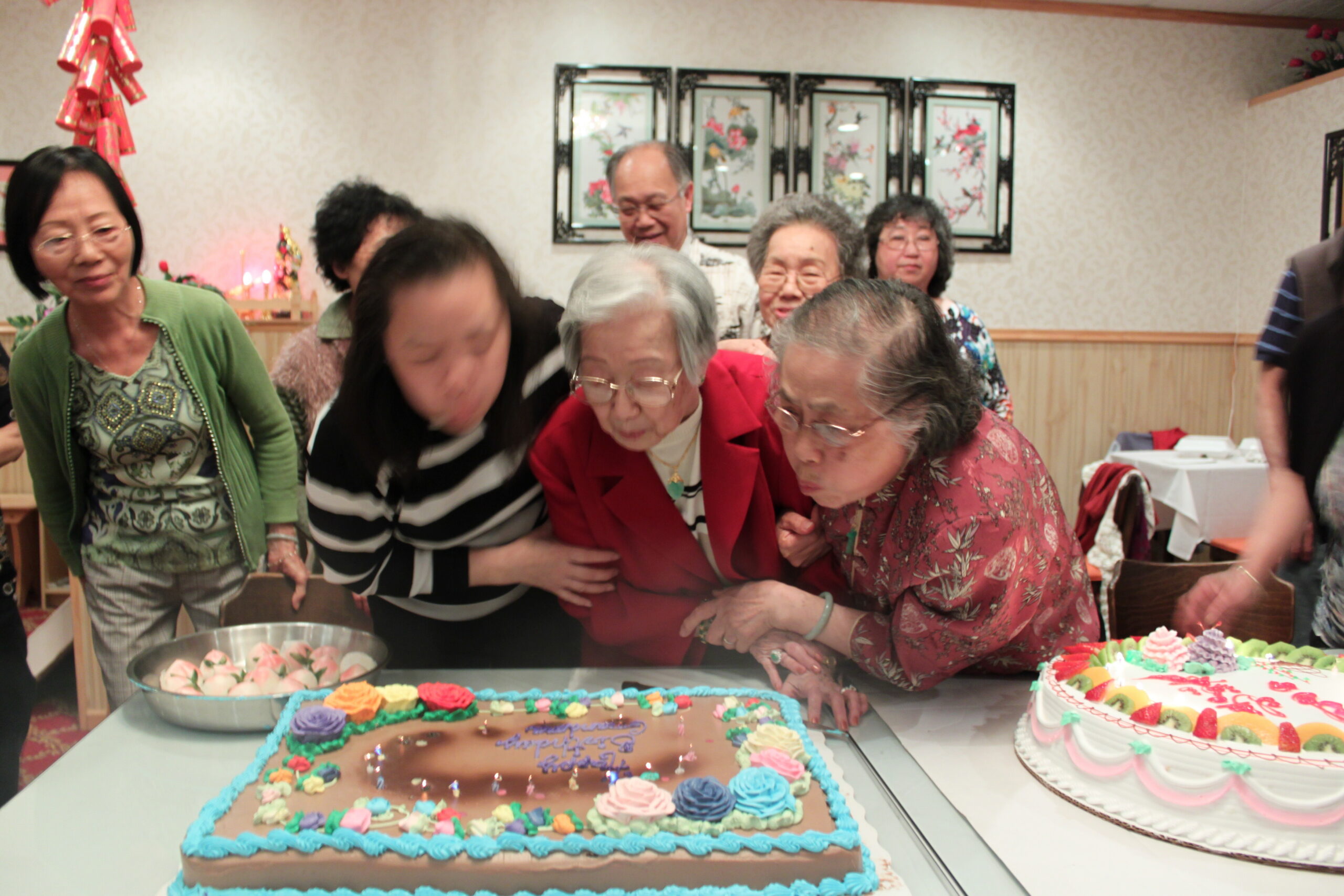 Group of elderly people gathered around a table to admire and cut a large decorated cake at a celebration, with cupcakes nearby in a bright room.