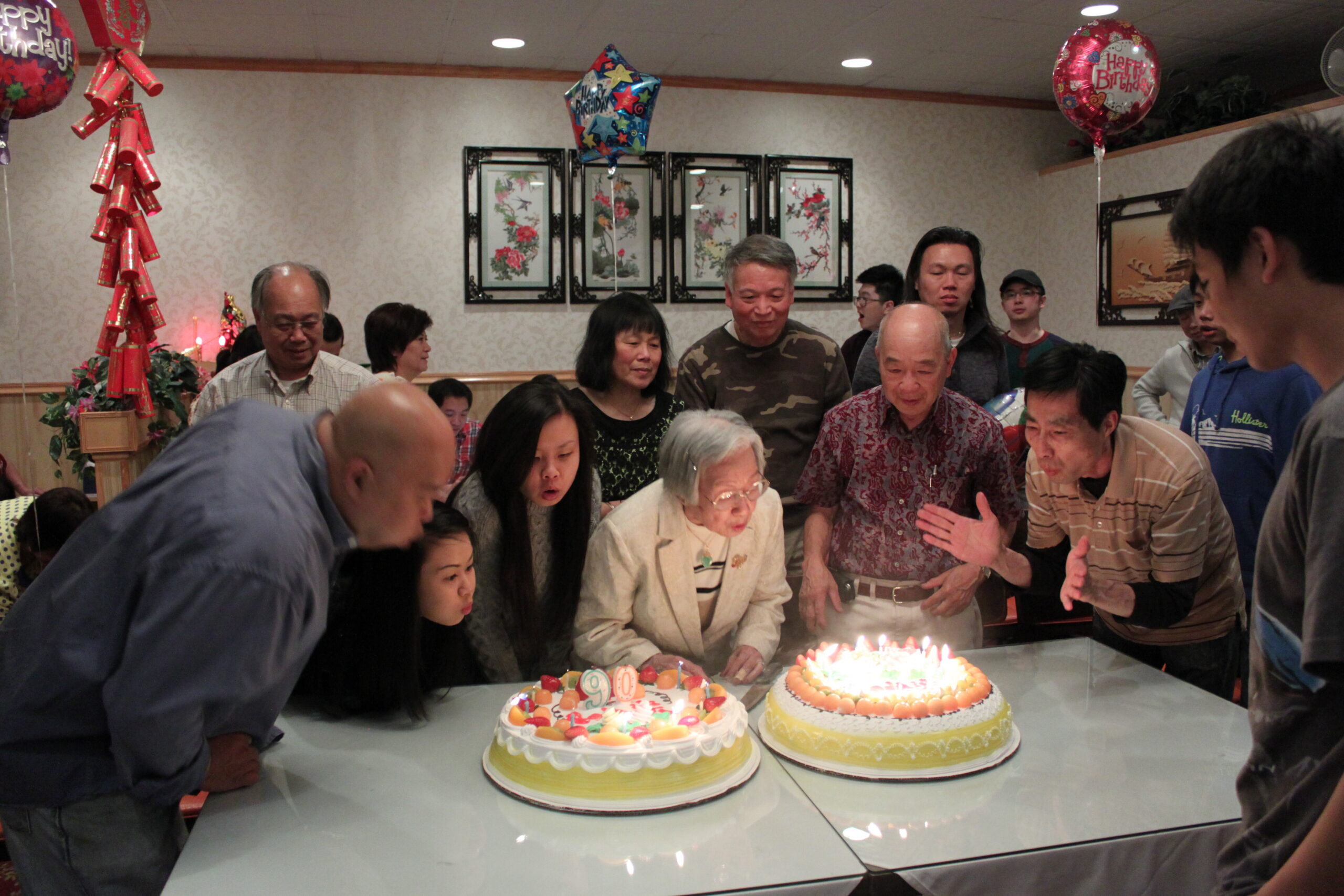A crowded birthday party with elderly and younger people gathered around two decorated cakes with lit candles in a banquet hall.