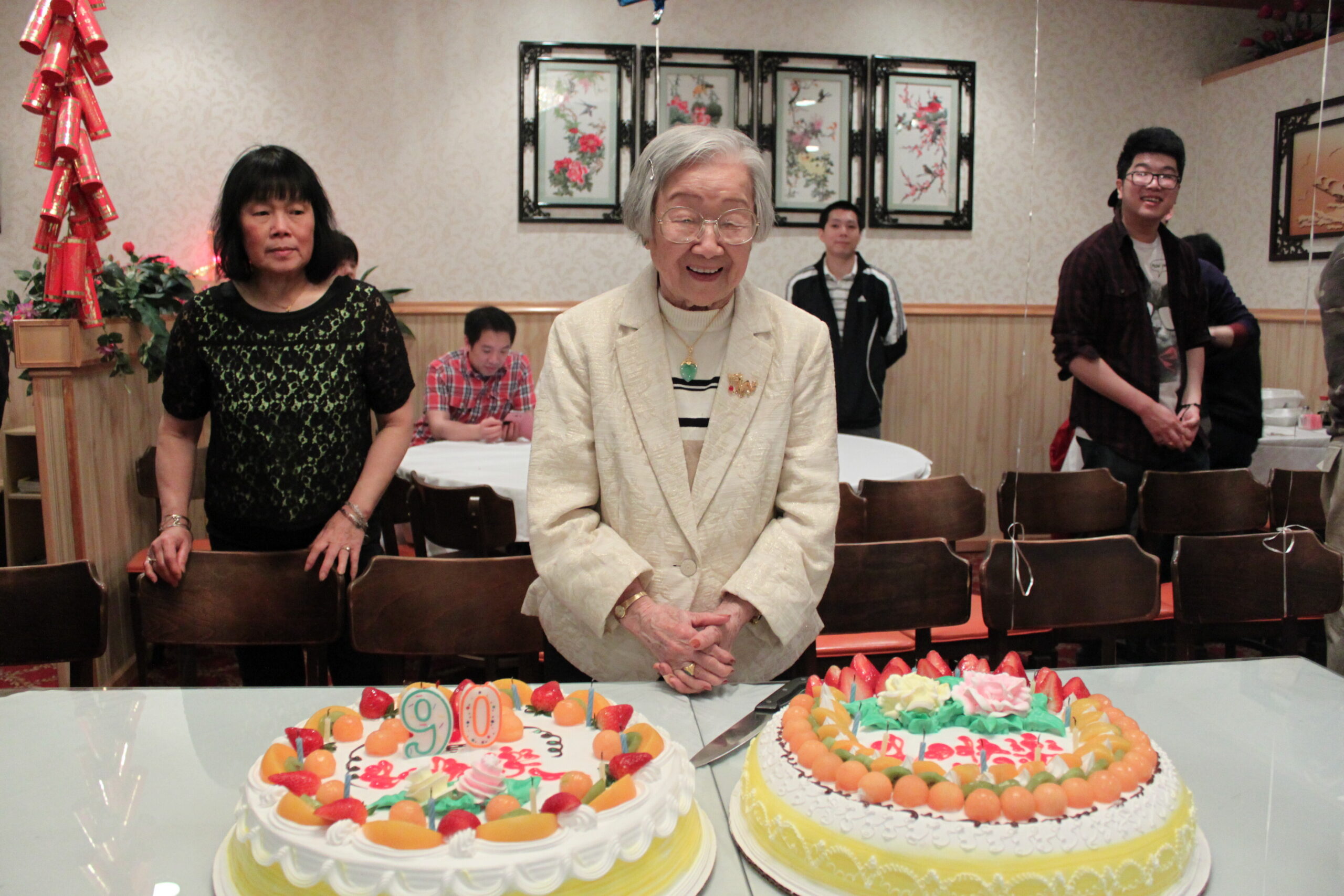 Elderly woman in a cream blazer smiles at two fruit-topped birthday cakes in front, with family behind her in a party setting.