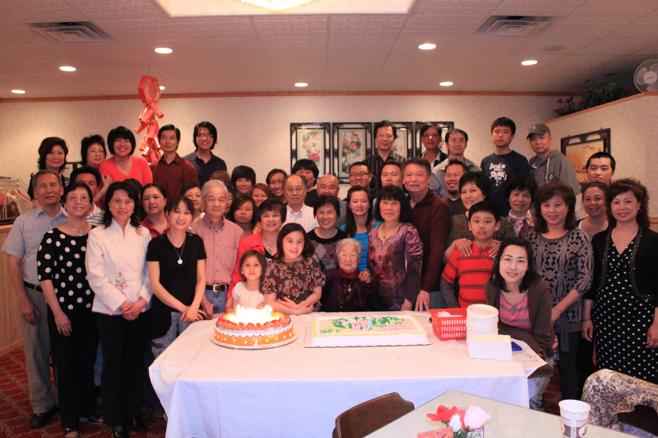 A large group of people gathered indoors around a table with two large birthday cakes, posing for a group photo at a celebration.