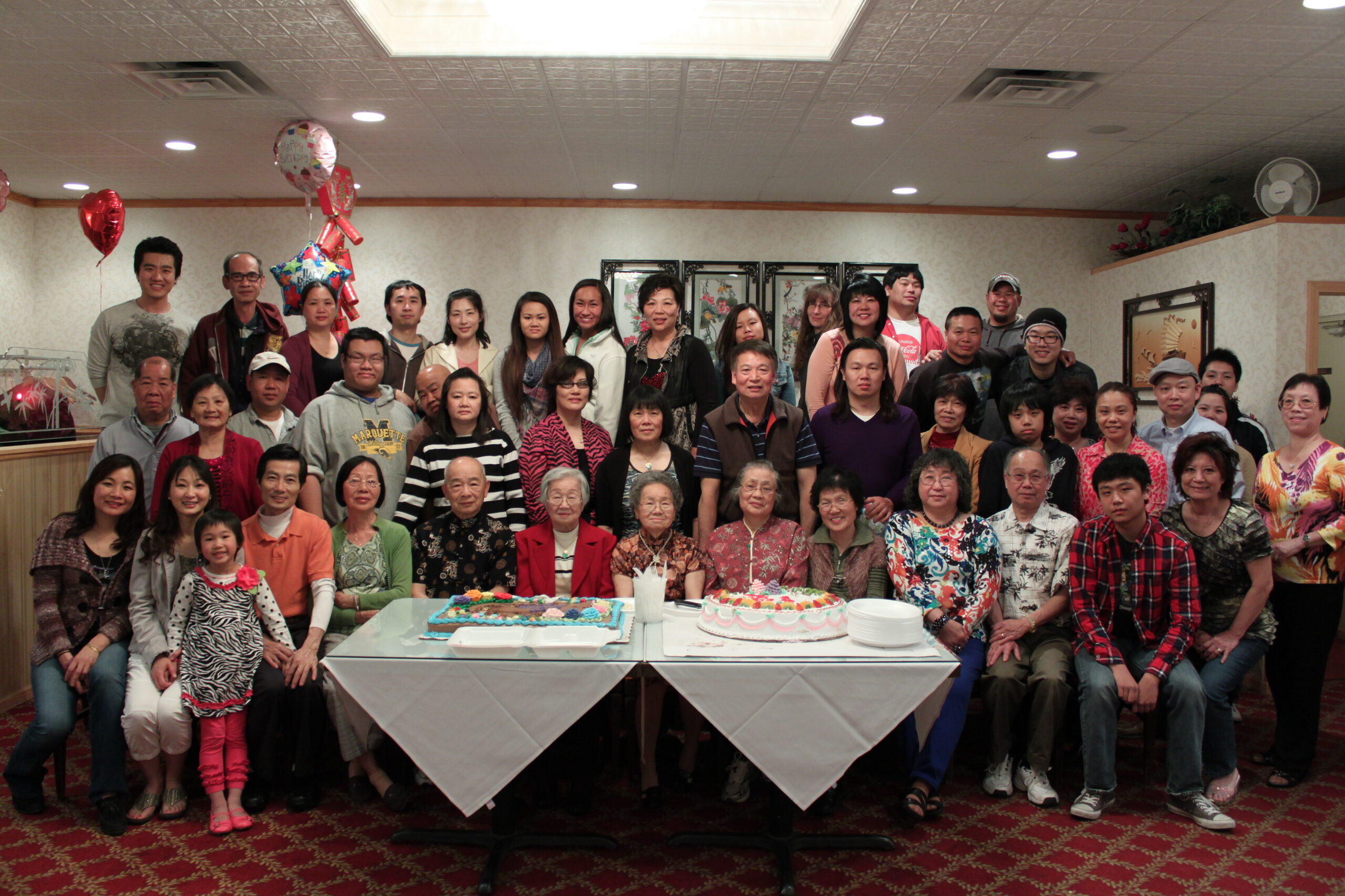 Large group of people posing for a birthday celebration in a banquet hall, with two cakes on a table in front and balloons in the background.
