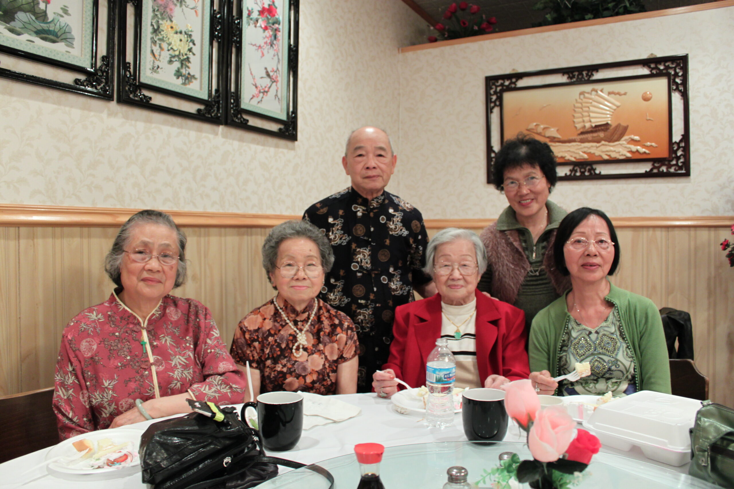 Five elderly Asian people posing for a group photo at a restaurant table with plates and drinks.