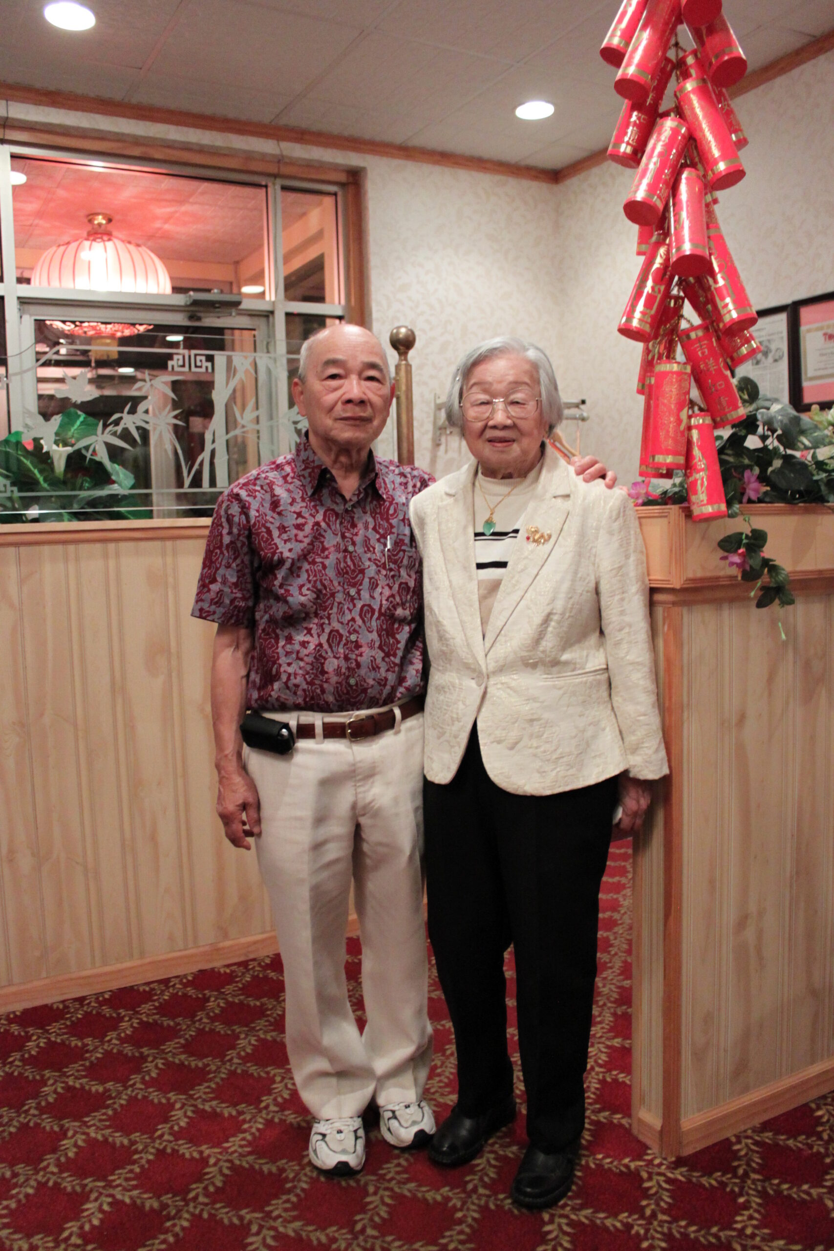 Two elderly Asian adults pose for a photo indoors next to a tall cluster of red decorative firecrackers hanging from the ceiling right of them.