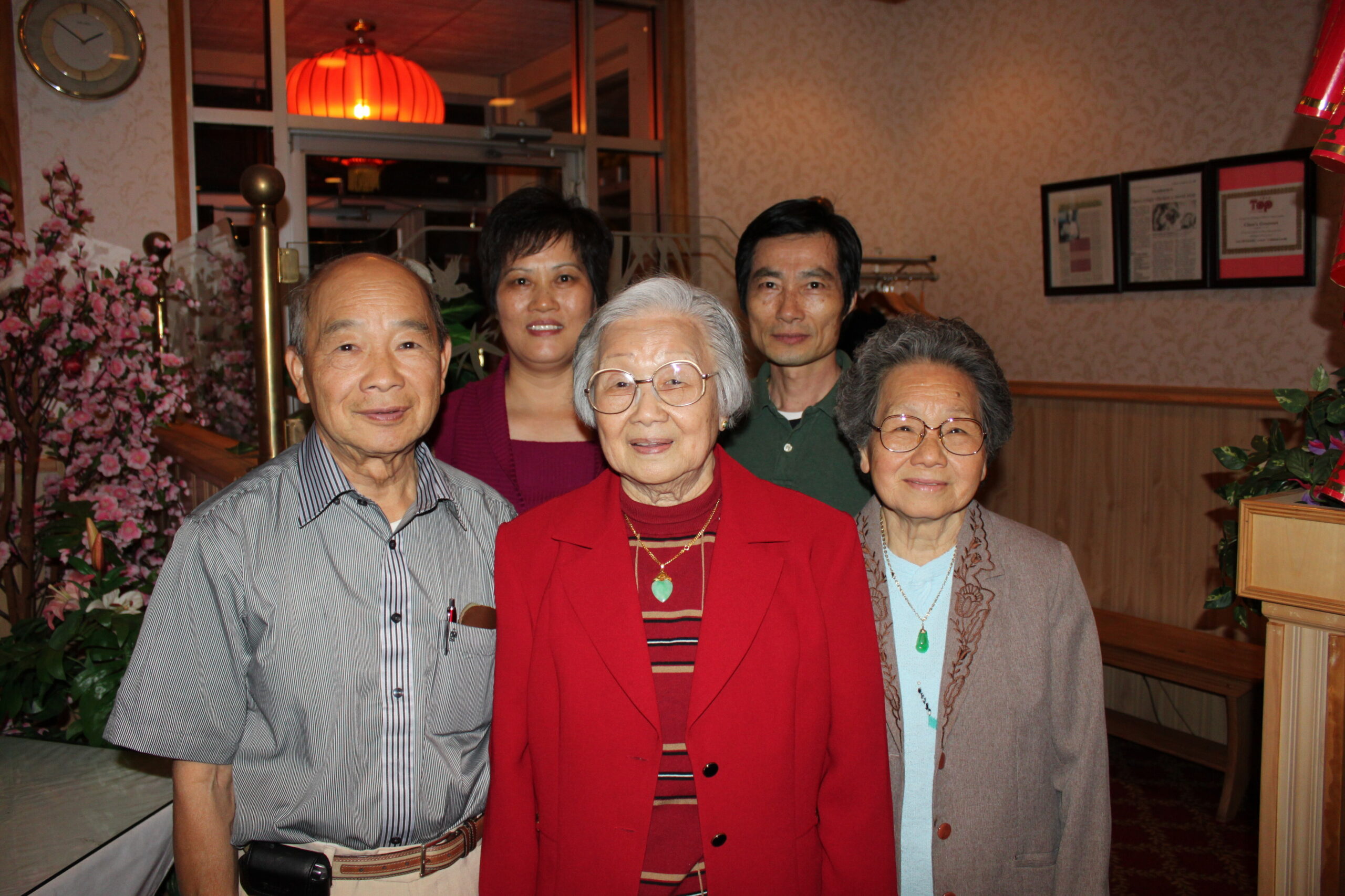 Group of five seniors posing for a photo in a restaurant, smiling at the camera with pink flowers in the background.