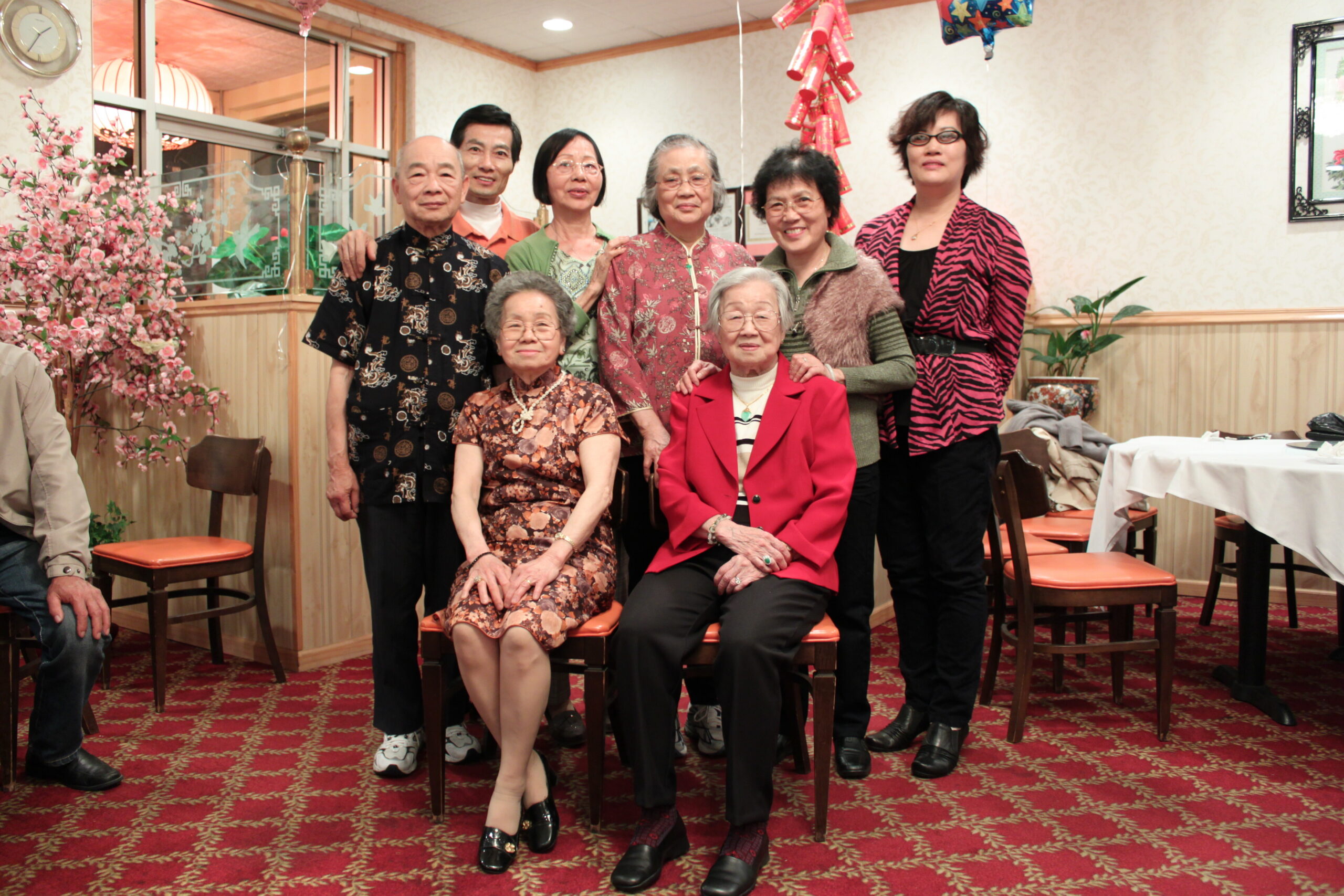 Group of nine Asian seniors posing for a photo in a restaurant-like banquet hall with pink cherry blossoms and festive decor behind them.