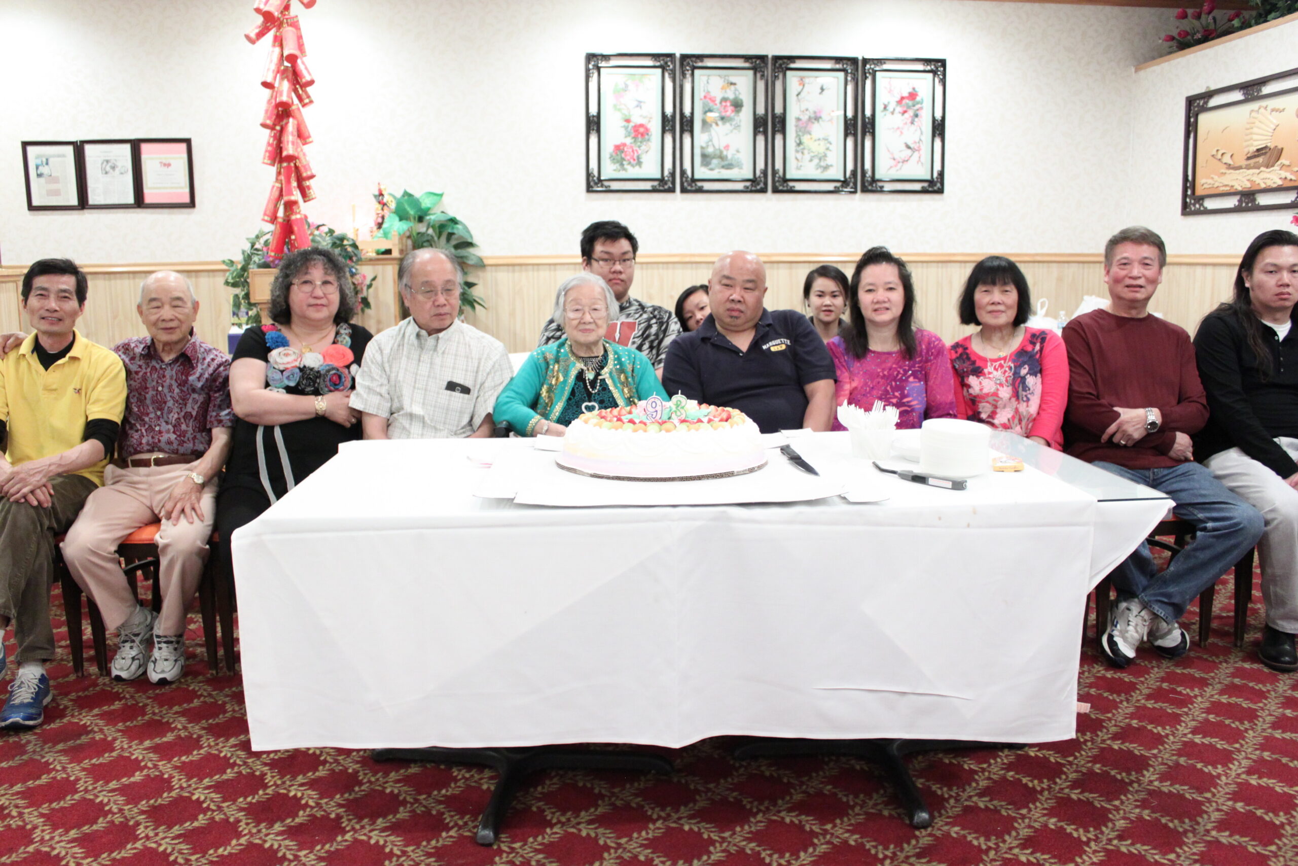 Group of people seated along a long table at a birthday celebration, cake with candles in the foreground and a decorative wall behind them.