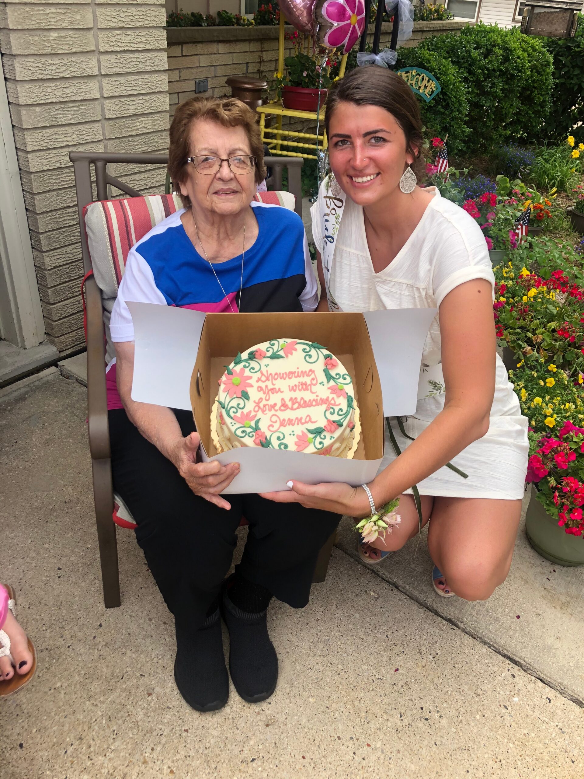 Older woman sitting on a porch chair next to a smiling young woman who holds a decorated birthday cake in a box; garden with flowers and balloons in the background.