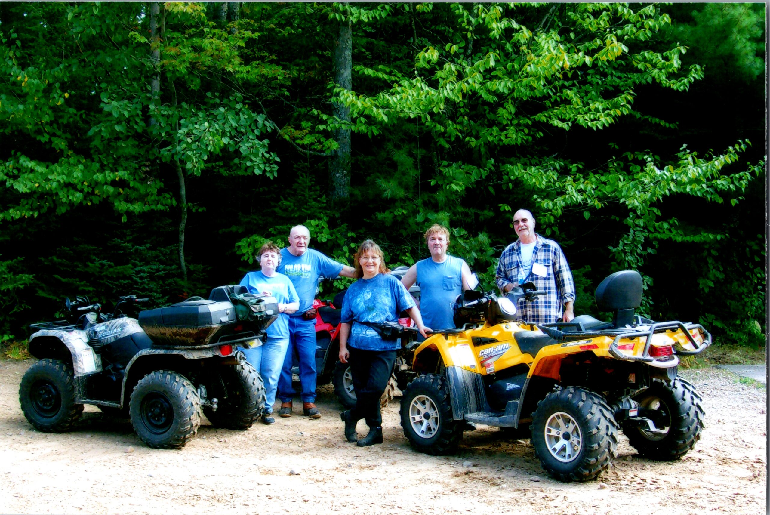 Group of people in blue shirts posing with all-terrain vehicles on a dirt path in a forested area.