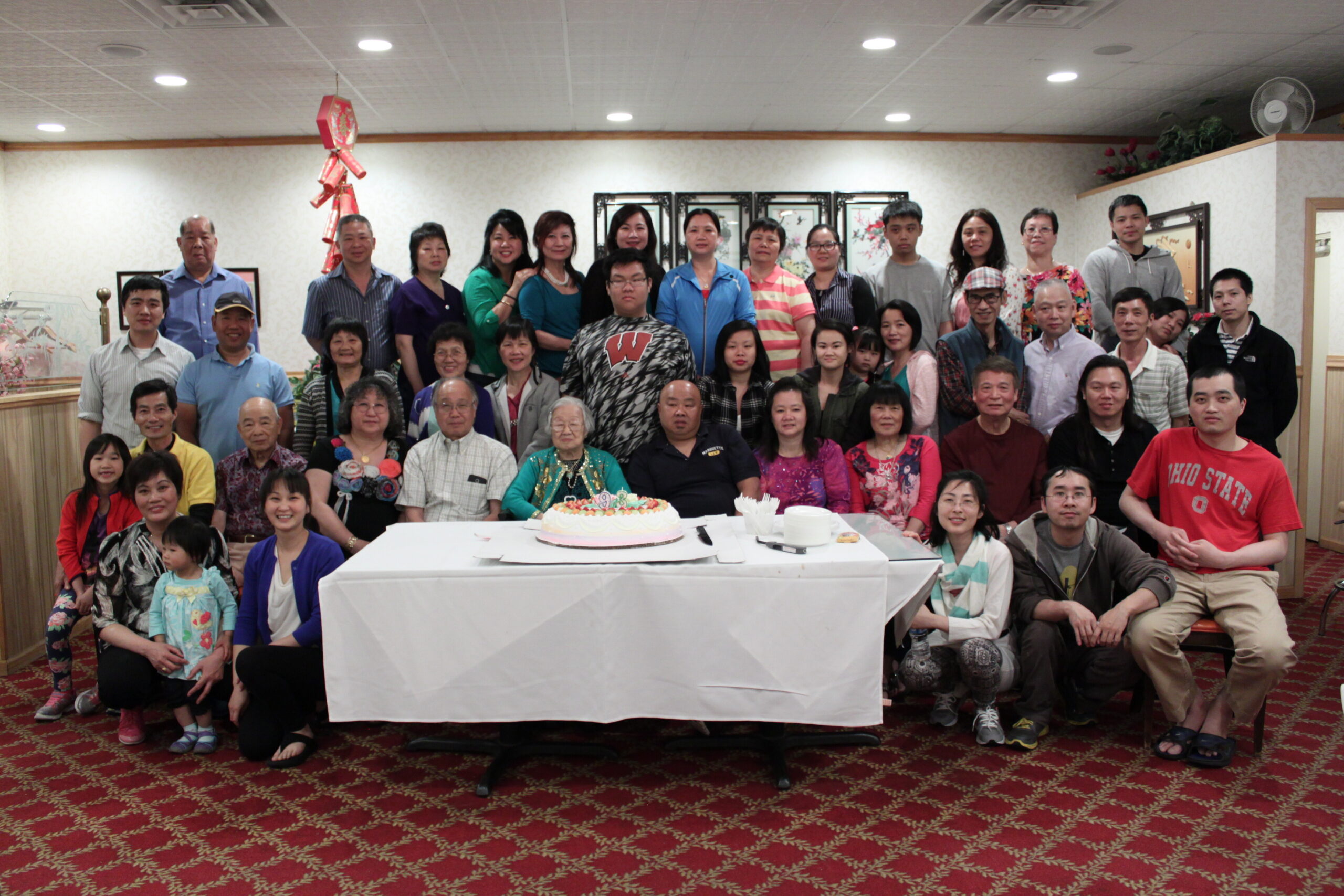 Large group of people posing around a white-covered table with a birthday cake in a banquet hall.