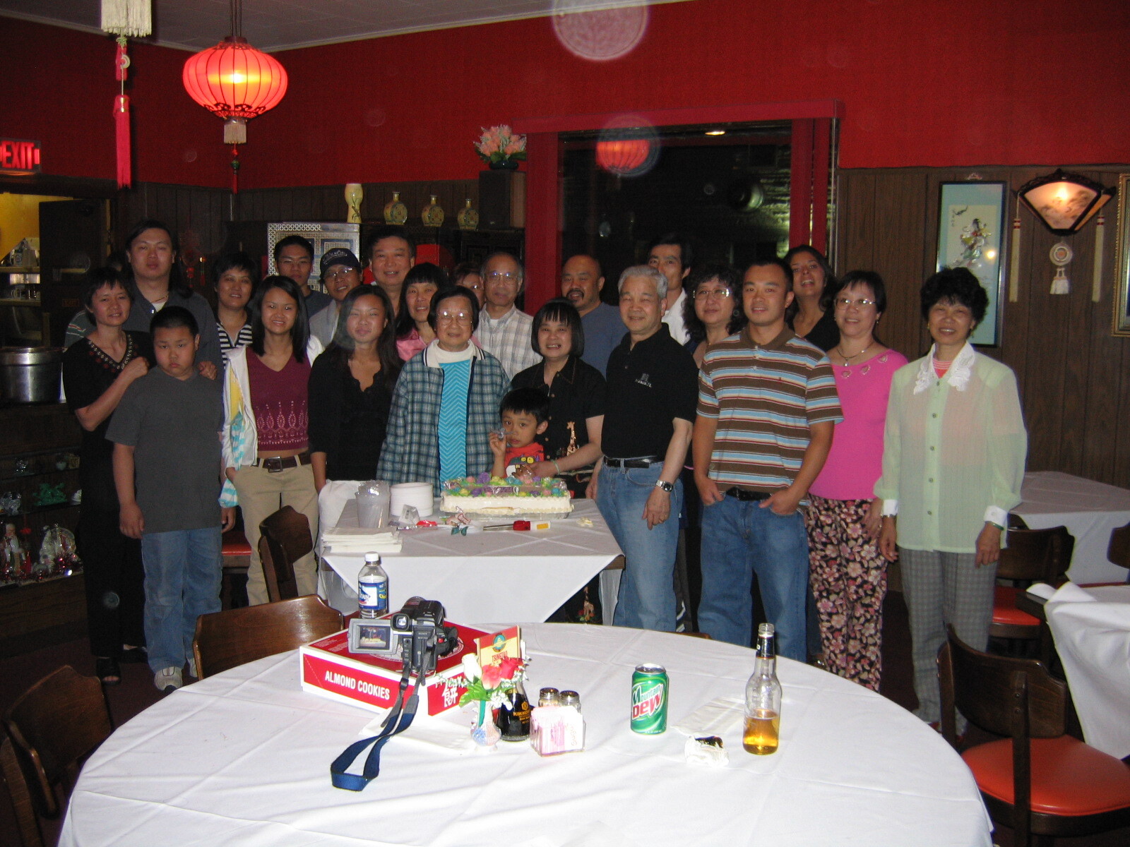 Group of people posing together in a restaurant for a celebration around a cake on a table with a child in the center and red lanterns overhead.