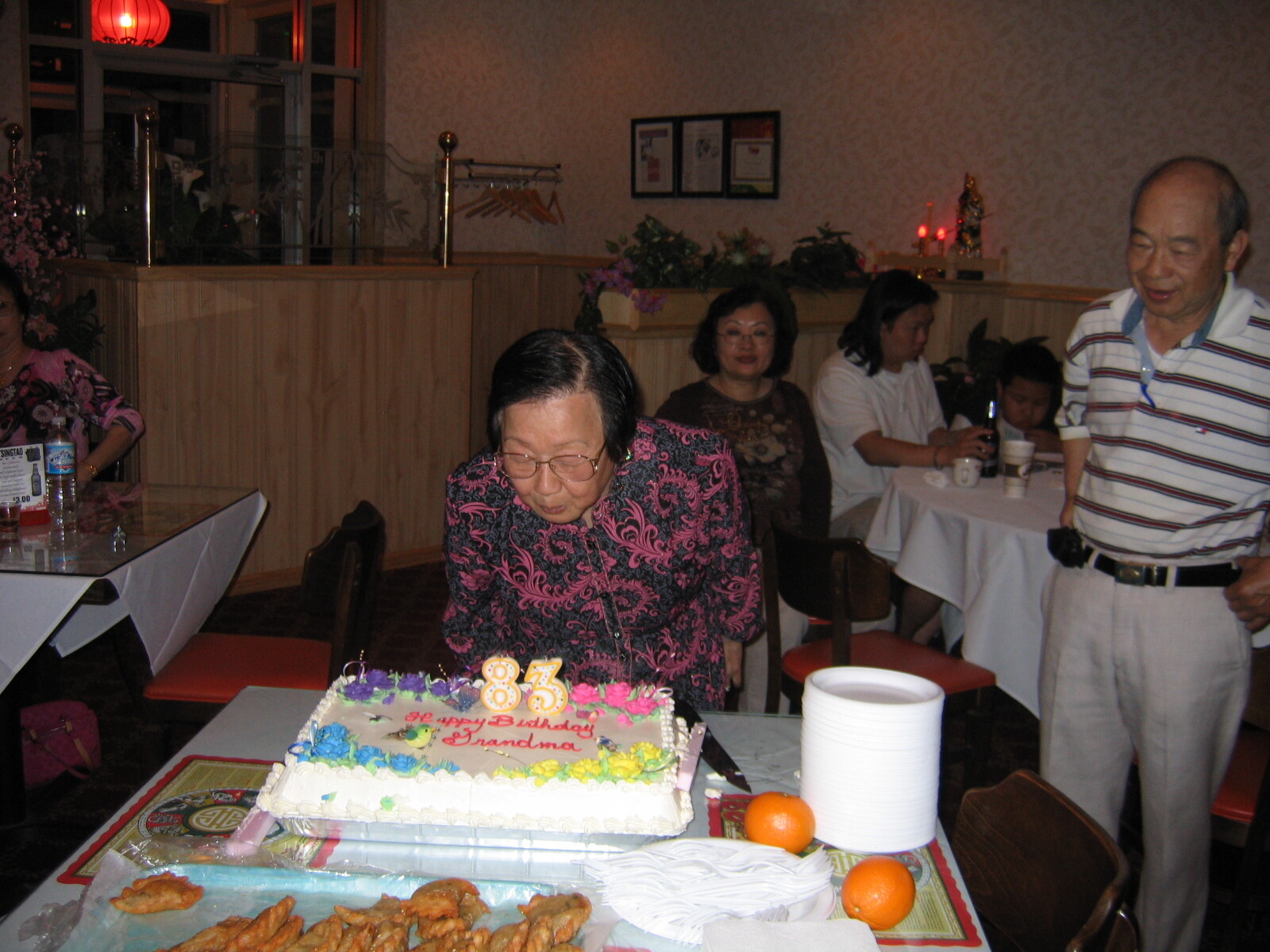 Older woman in pink patterned blouse leaning toward a birthday cake with lit 83 candles; friends sit and chat in a restaurant setting behind her.
