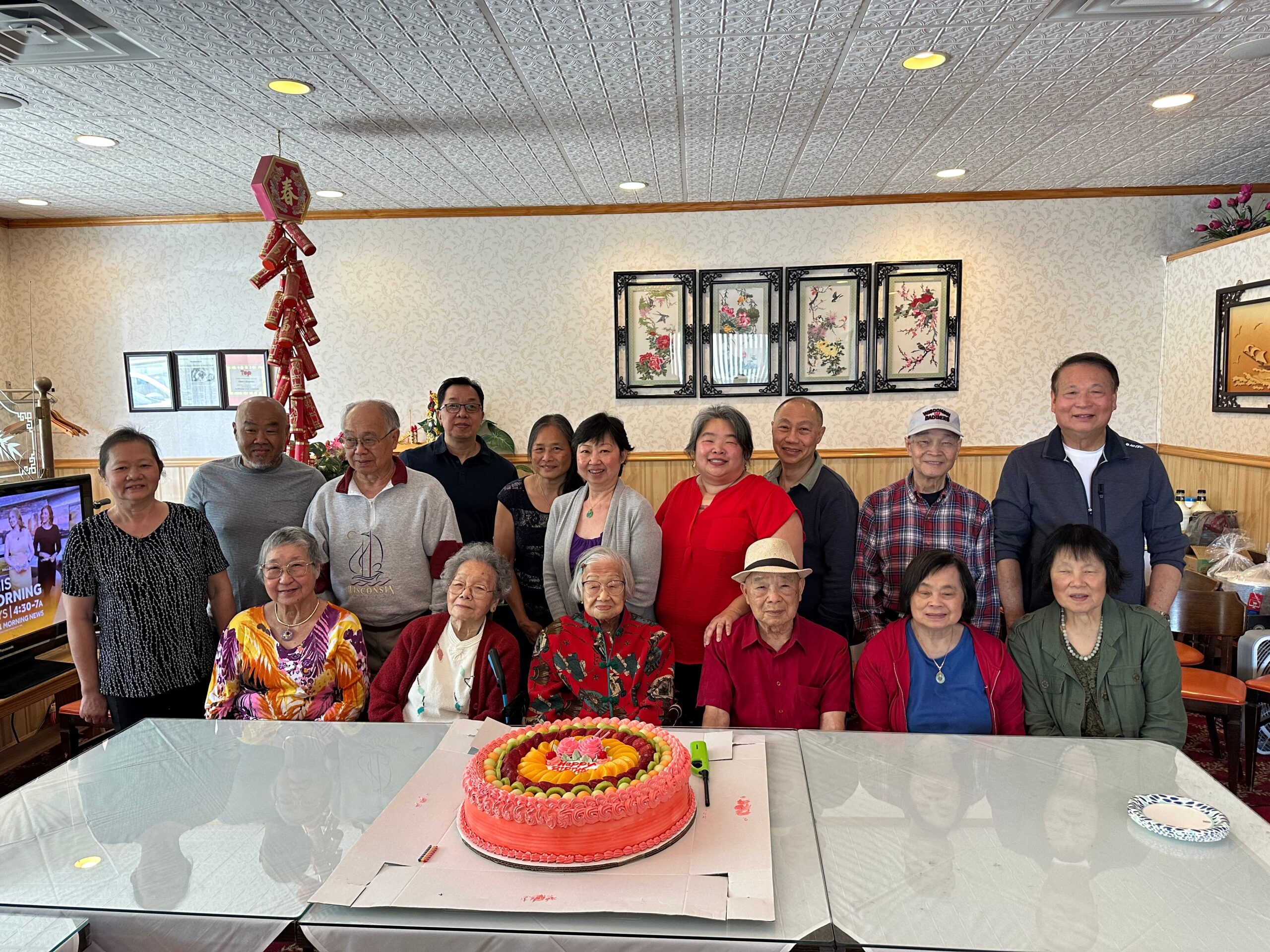 Group of smiling seniors and others posing behind a large pink birthday cake in a restaurant setting.