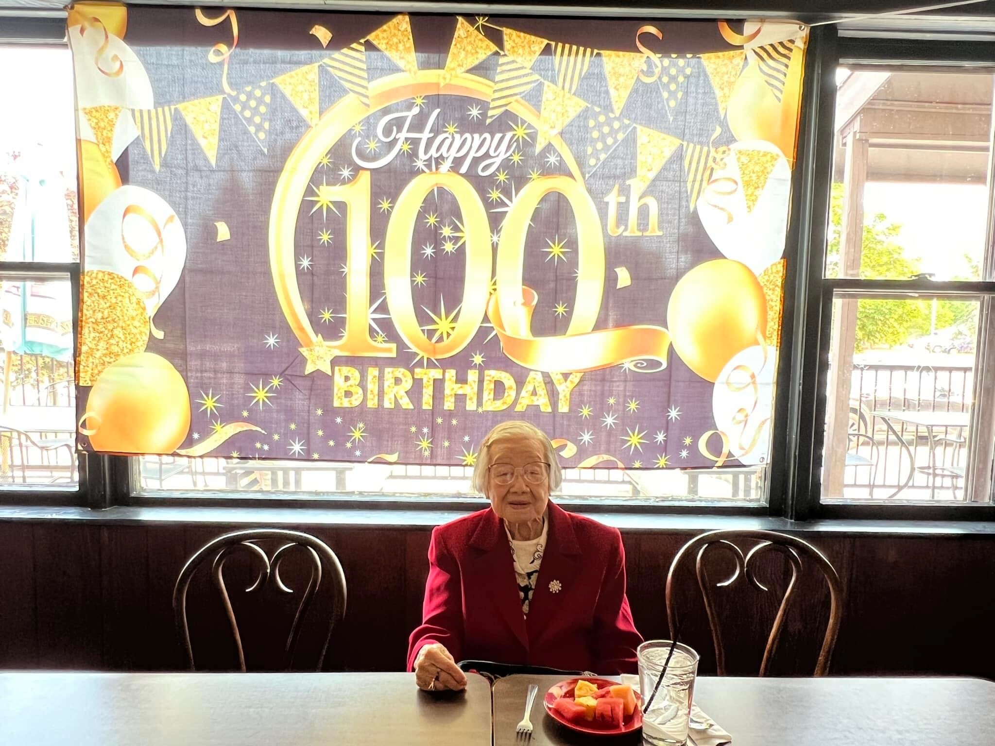 Elderly woman in a red blazer sits at a table with a fruit plate, in front of a purple-gold 'Happy 100th Birthday' banner with balloons.