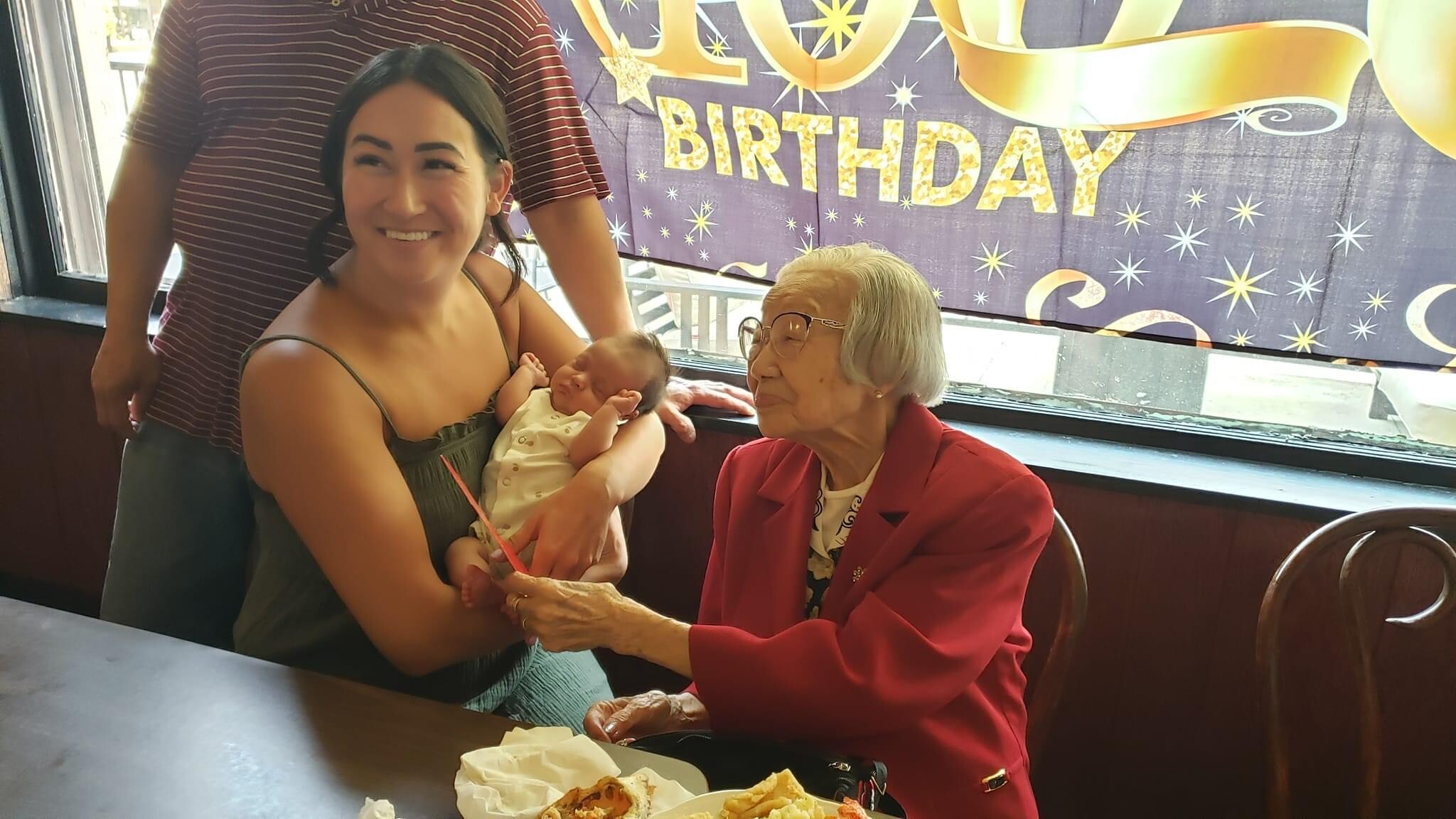 Woman holds a smiling baby while an elderly lady in a red blazer sits nearby at a birthday party in a restaurant.