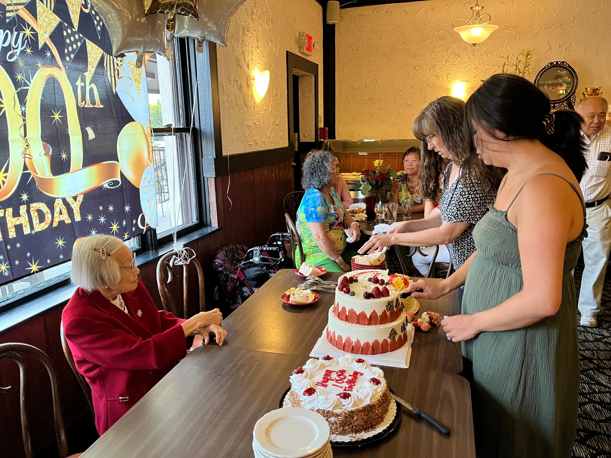 Older woman in a red jacket sits at a table as others stand around, birthday cake being served nearby in a cozy restaurant.