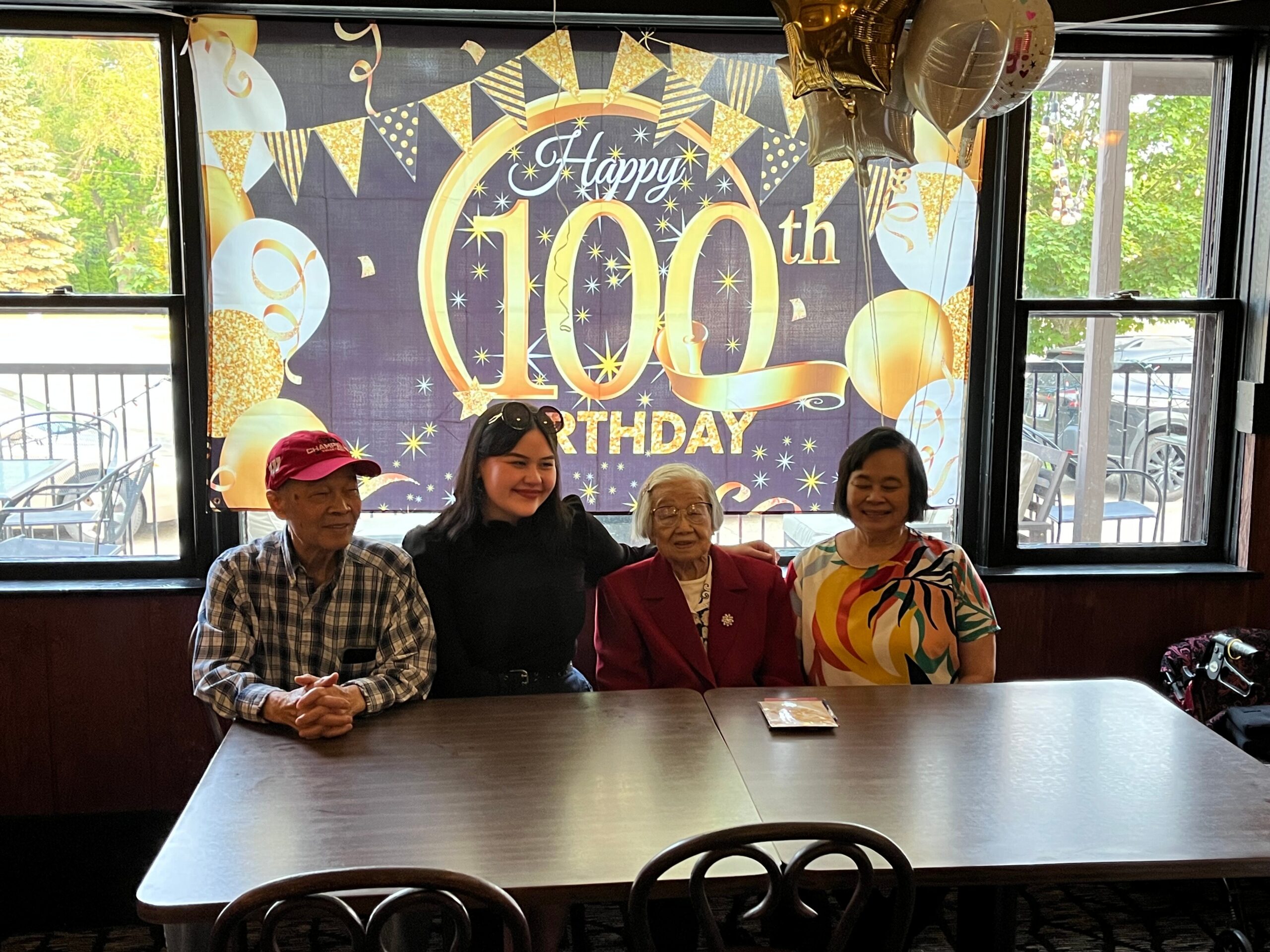 Group of five seniors posing at a 100th birthday party with a festive banner and balloons in the background.