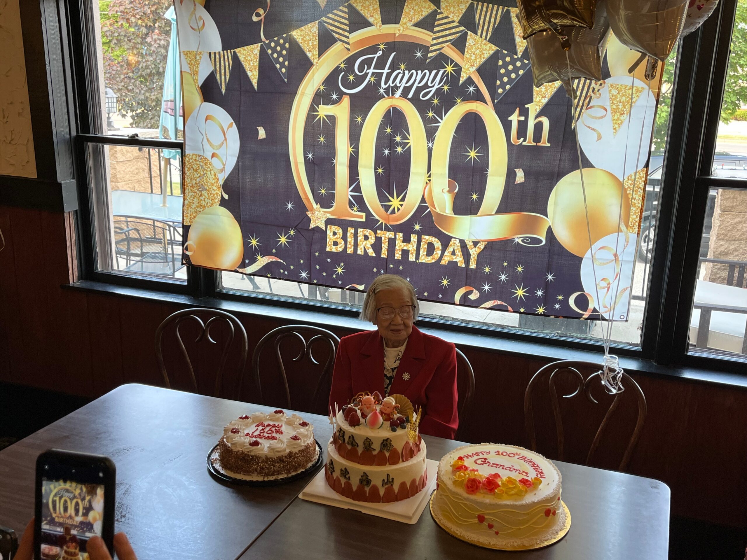 Elderly woman sitting at a table with three birthday cakes in front of a large glittery 'Happy 100th Birthday' banner and balloons outside a window.