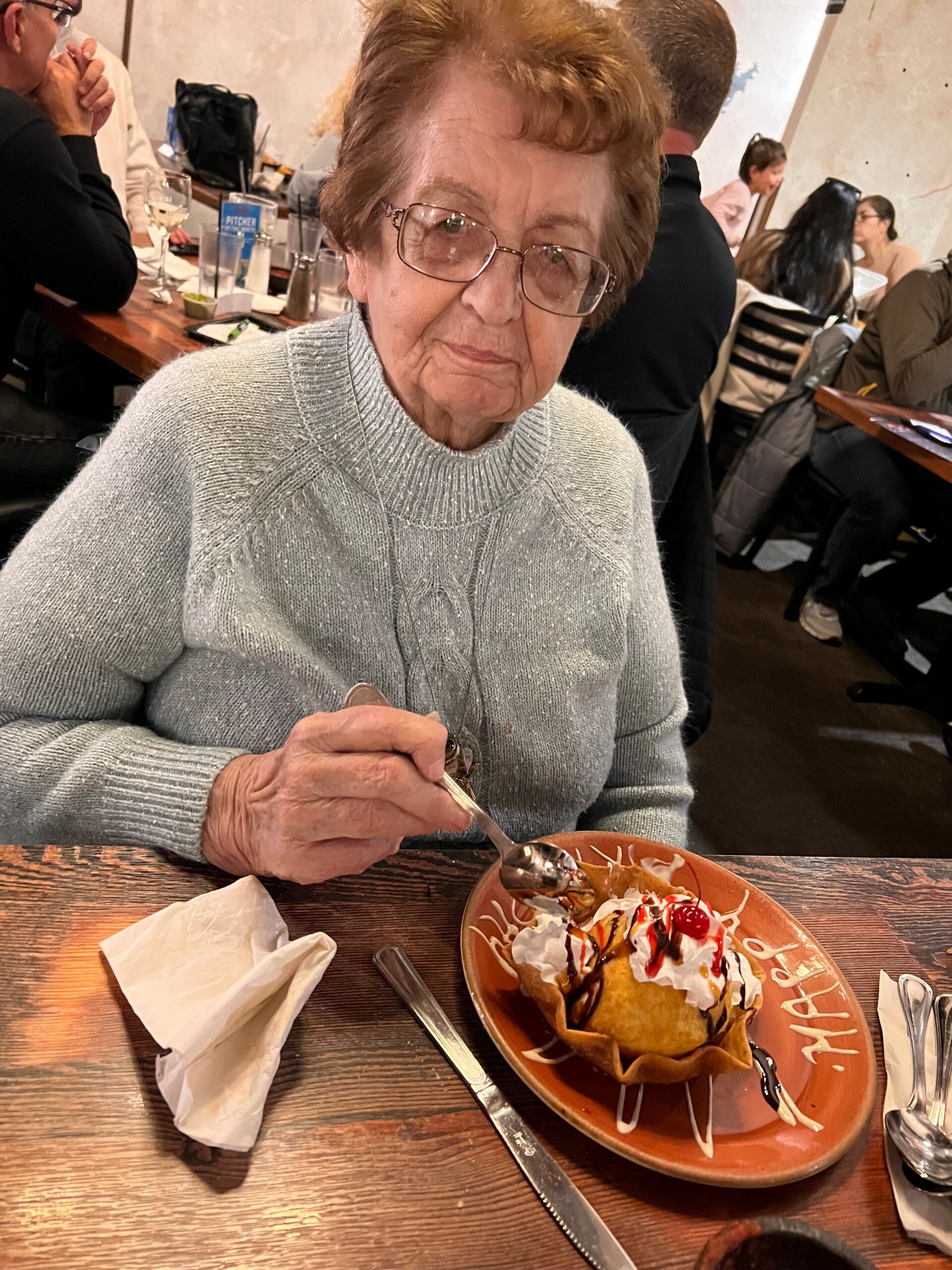 Older woman with glasses in a gray sweater at a restaurant, holding a spoon over a dessert topped with whipped cream and cherries on a brown plate.