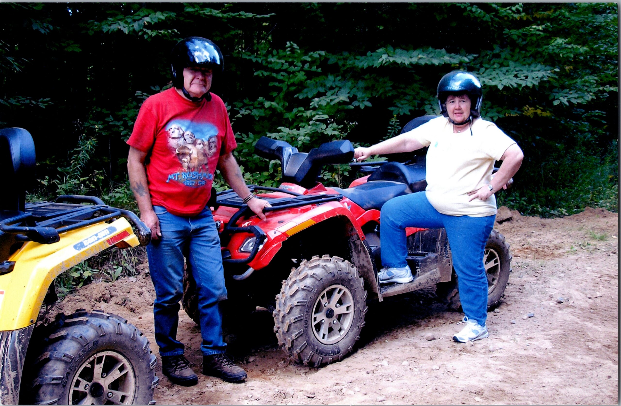 Two adults wearing helmets lean on red and yellow ATVs on a dirt trail with green foliage behind them.