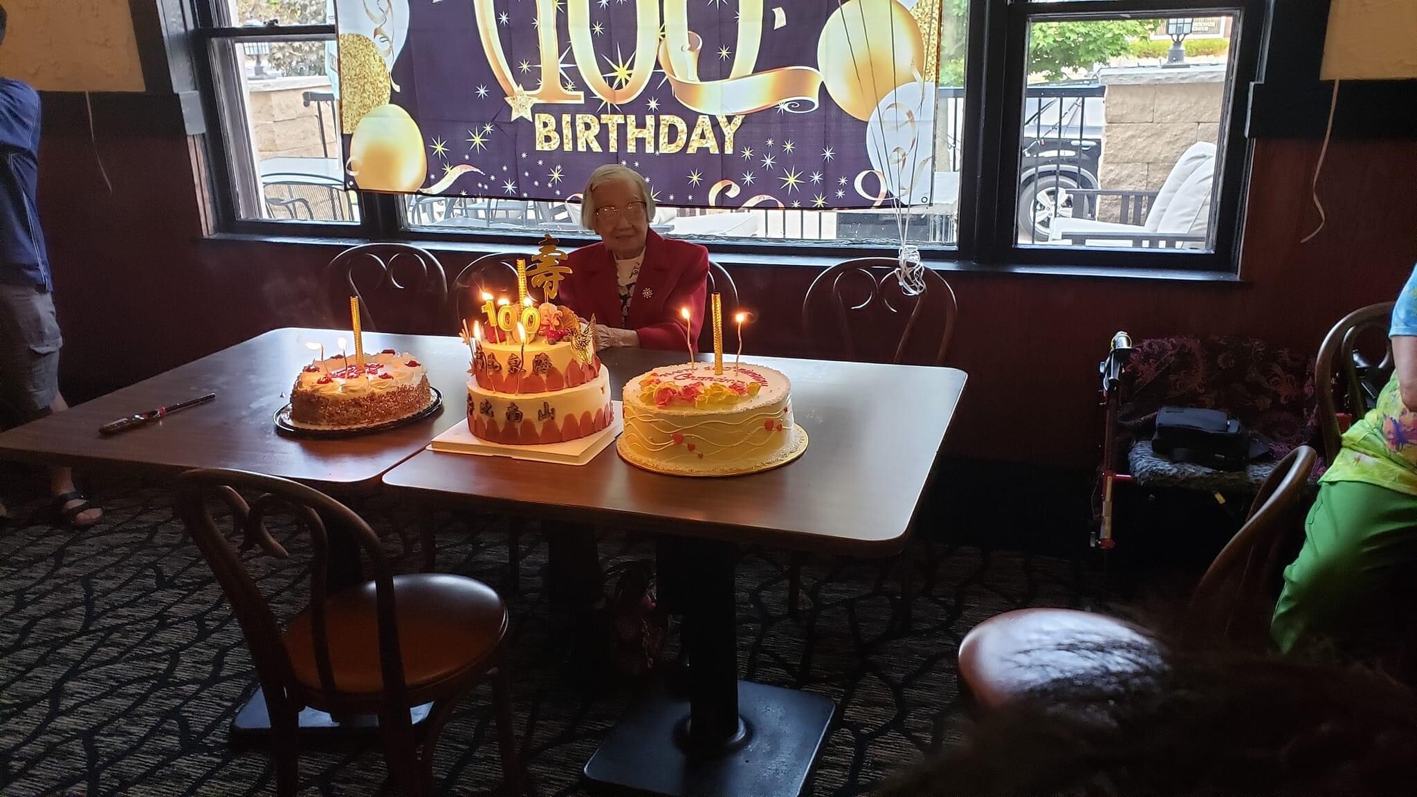 An elderly woman in a red blazer seated at a table with two decorated birthday cakes and lit candles during a birthday celebration in a restaurant.