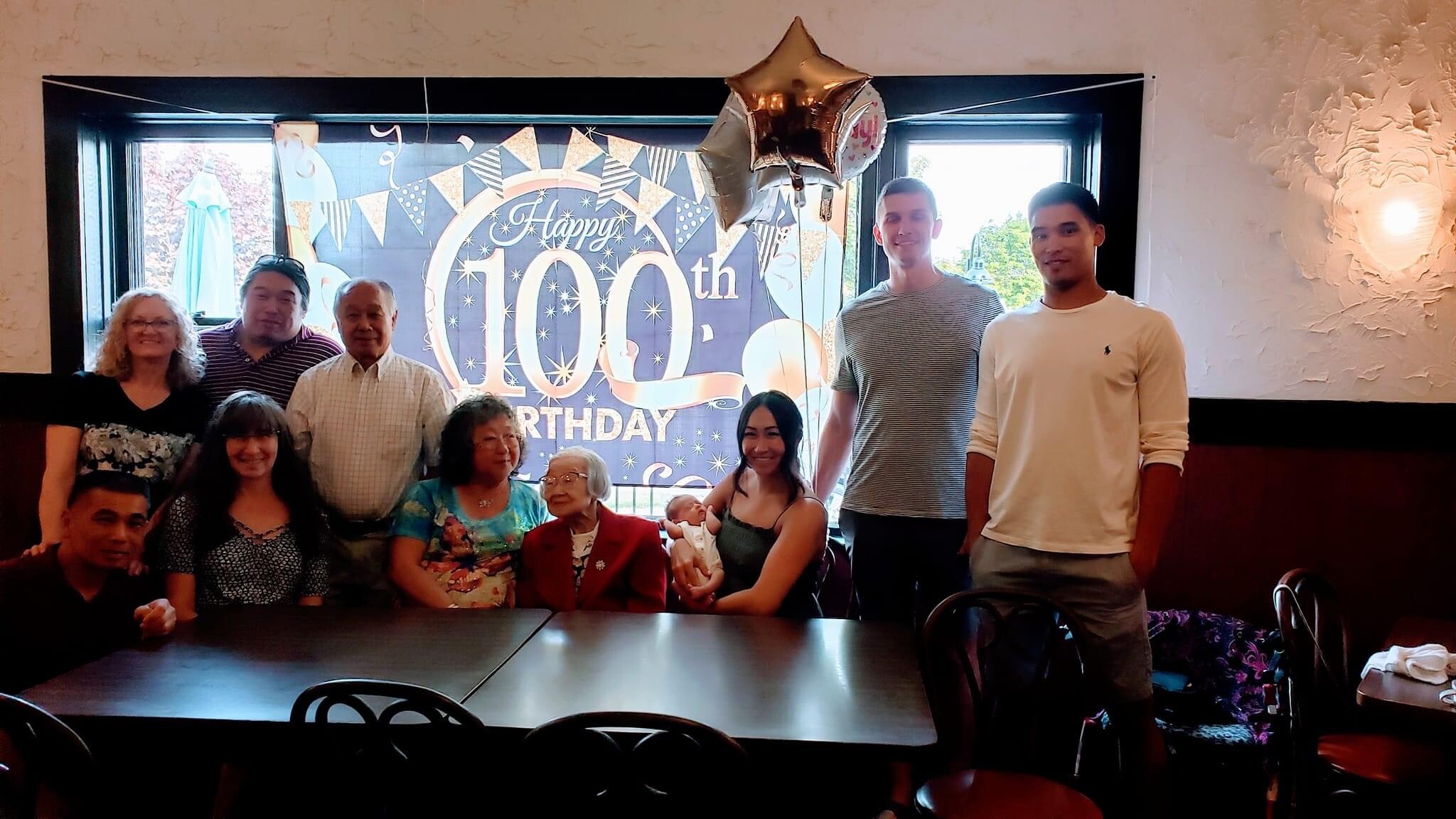 Group of family and friends posing indoors in front of a 'Happy 100th Birthday' banner with balloons, celebrating a milestone event.