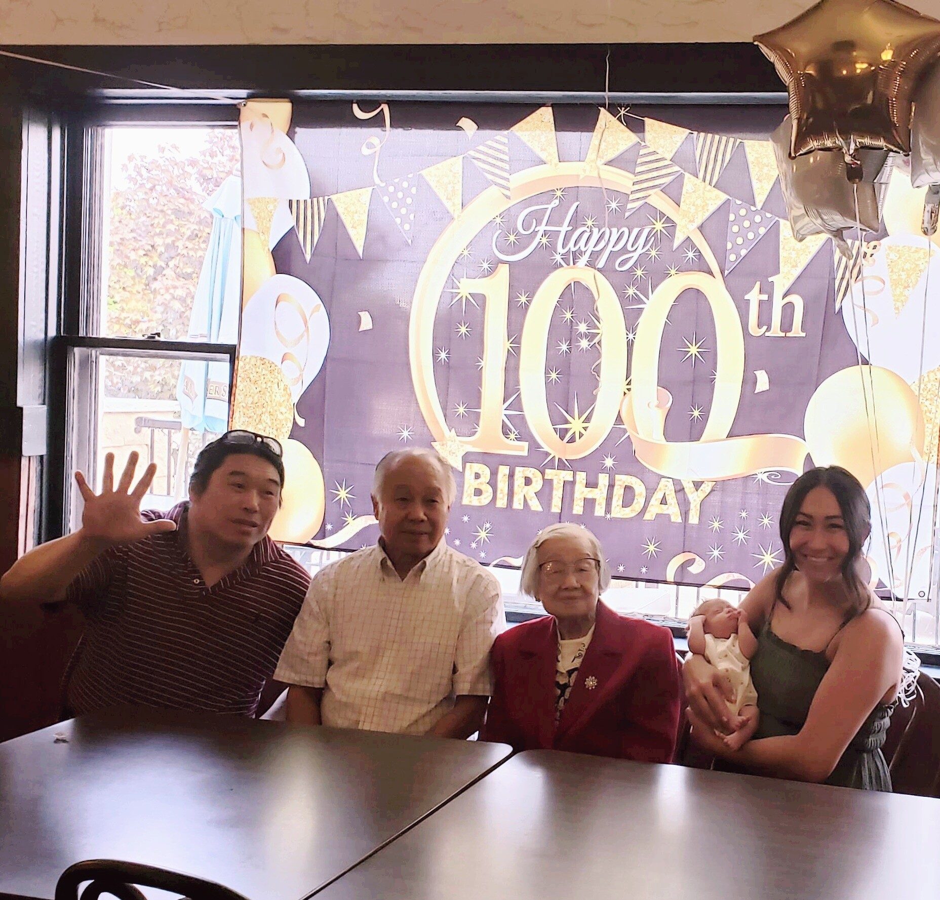 Group of relatives posing at a birthday party in front of a large 'Happy 100th Birthday' banner.
