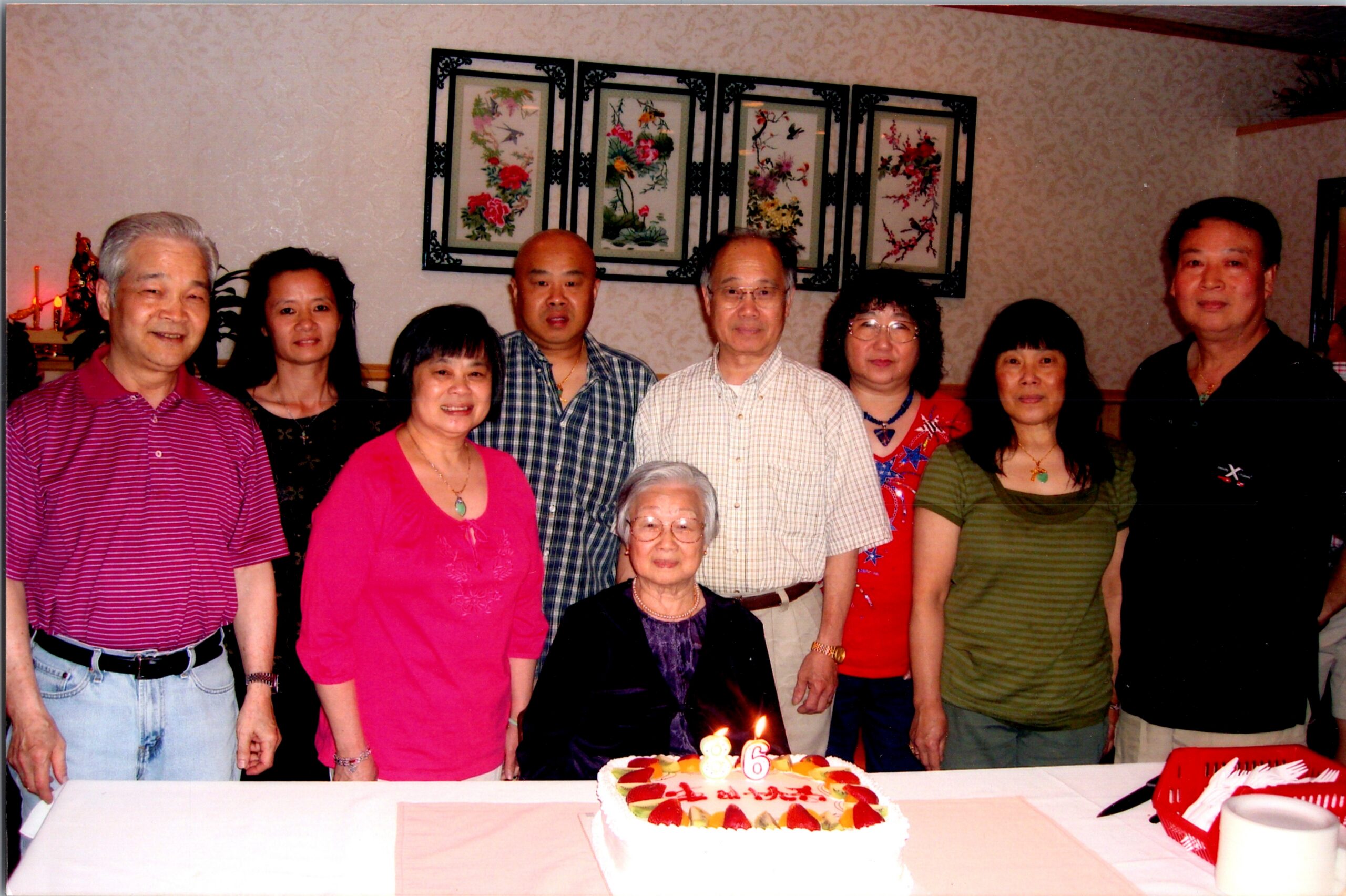 A group of adults posing indoors around a decorated birthday cake with candles on a table in front of them.