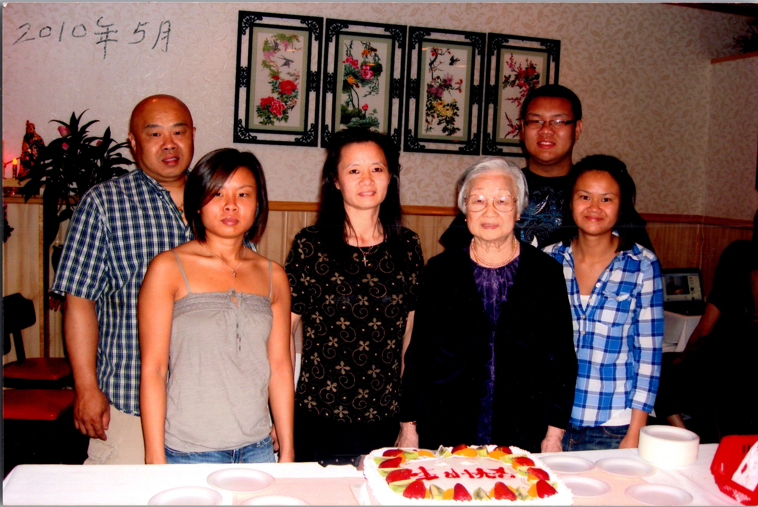 Six adults posing together at a birthday party, with a fruit-topped cake in front on a table in a dining room.