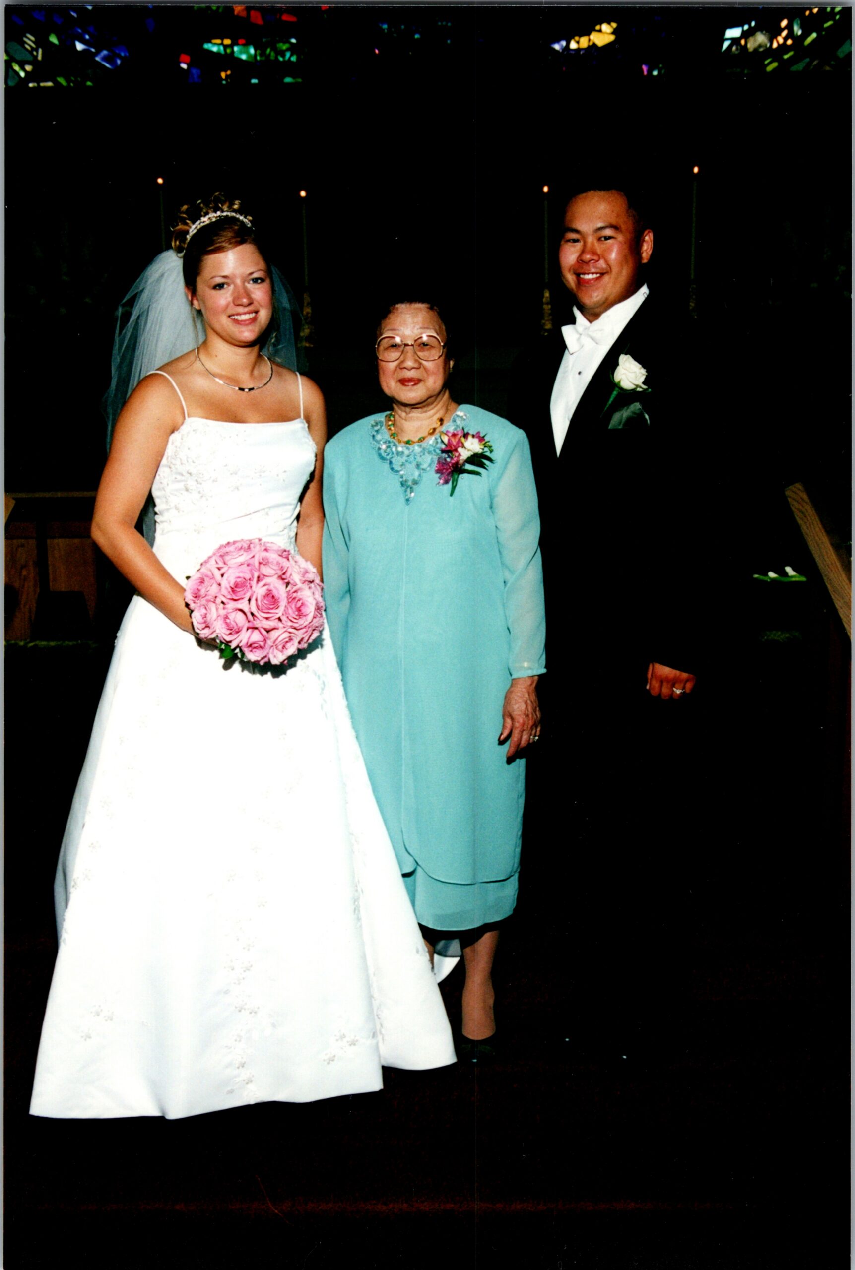 Bride in a white wedding gown holds a pink bouquet, standing with an older woman in teal and a smiling groom in a tuxedo.