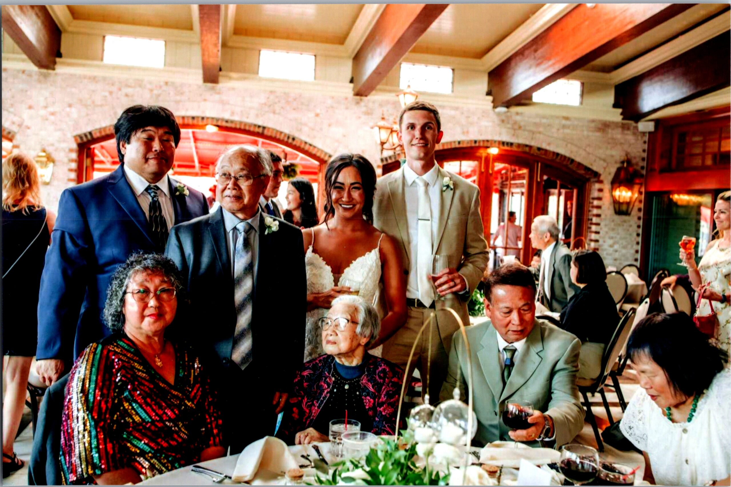 Bride in a white gown posing with two men at a wedding reception in a brick-walled, rustic venue with guests seated around tables.