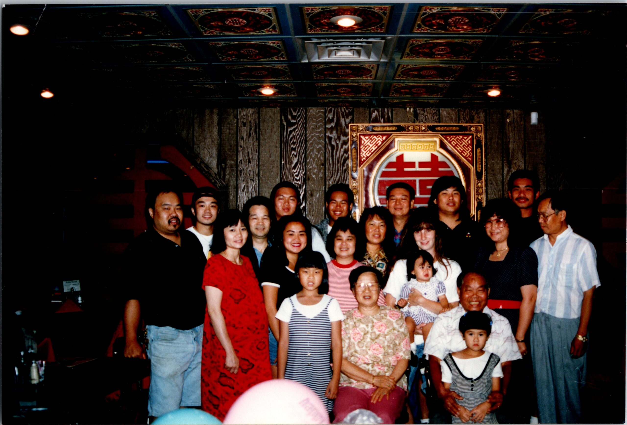 Group photo of about 25 people, adults and children, posing together in a dim venue with ornate ceiling tiles and a large decorative emblem behind them.