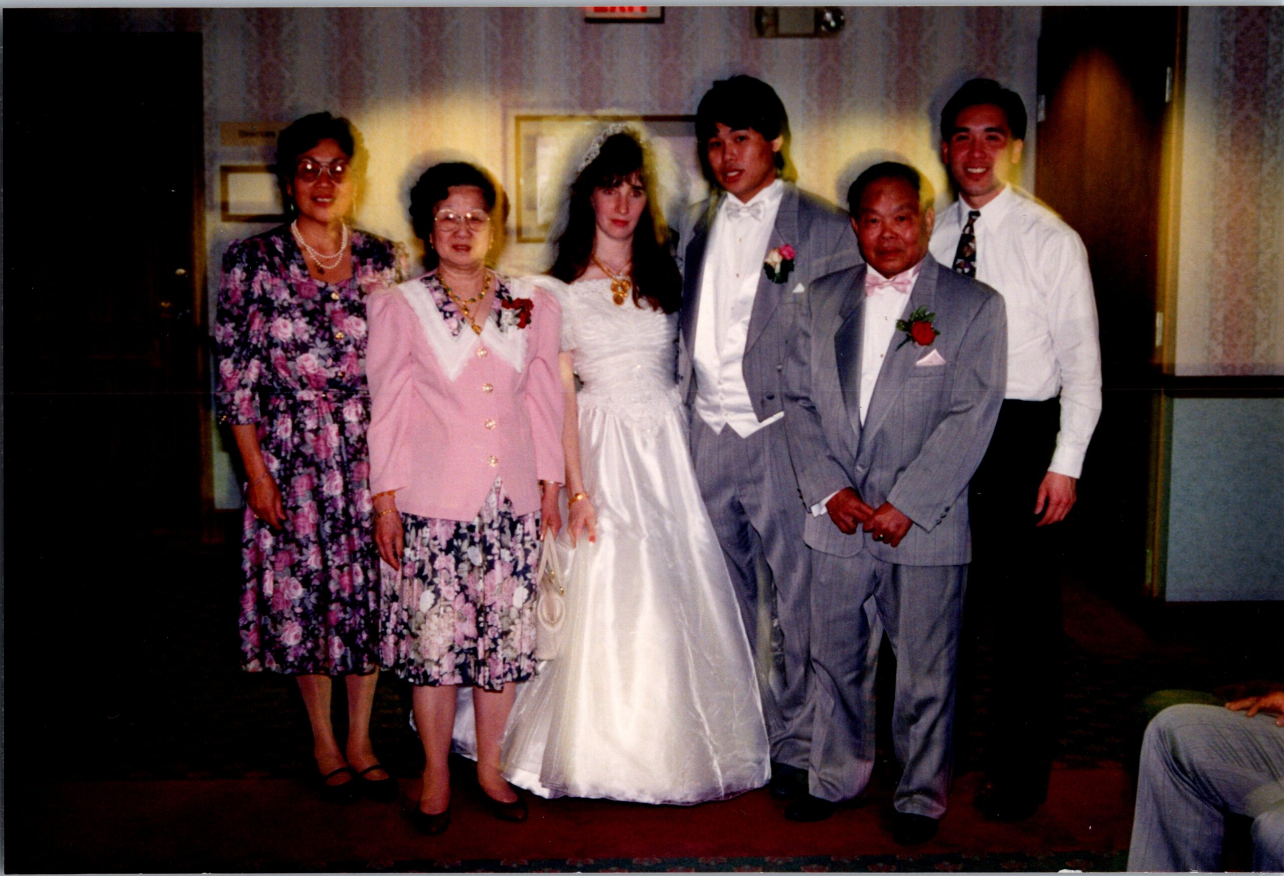 Bride in a white wedding gown stands with her groom in a light suit and several family members in dresses and suits at a wedding reception.