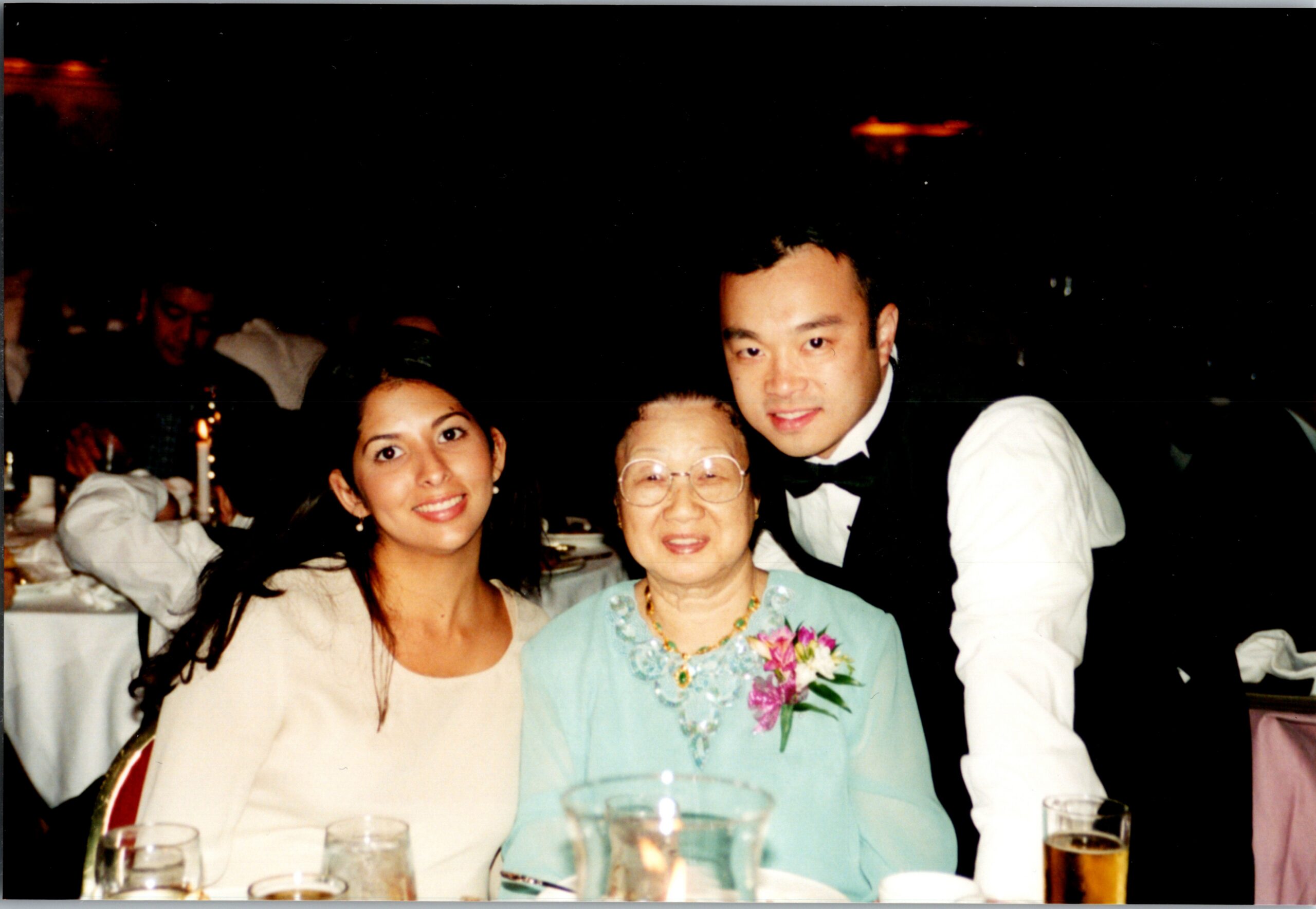 Three people pose at a banquet table: a younger woman left, an elderly woman center in turquoise with a corsage, and a man in formal attire on the right.