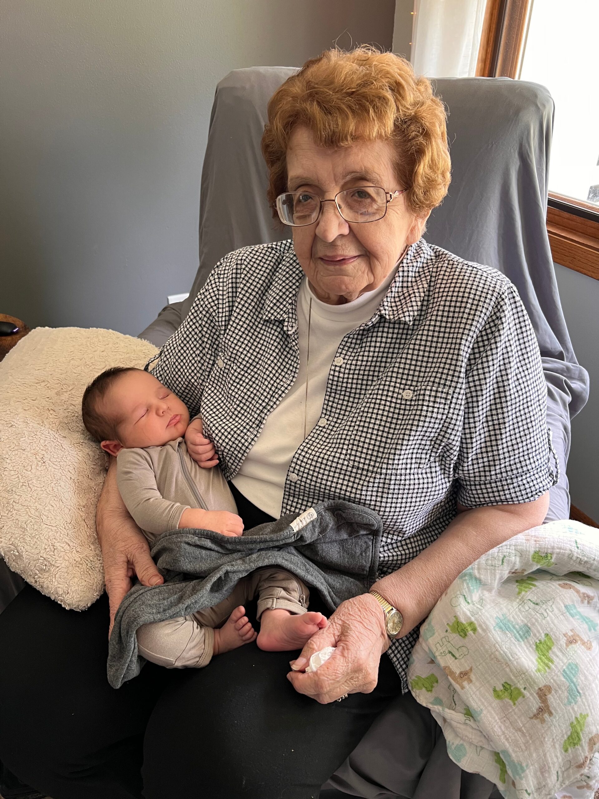 Older woman with short curly hair and glasses sits in a chair, cradling a sleeping newborn wrapped in a gray blanket.