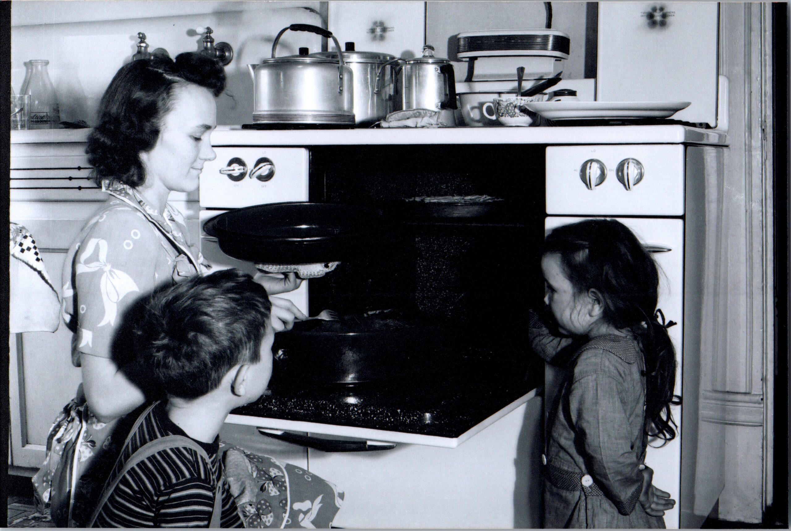Woman in an apron helps two children at a vintage stovetop with pots and dishes on the counter behind them in a mid-century kitchen.
