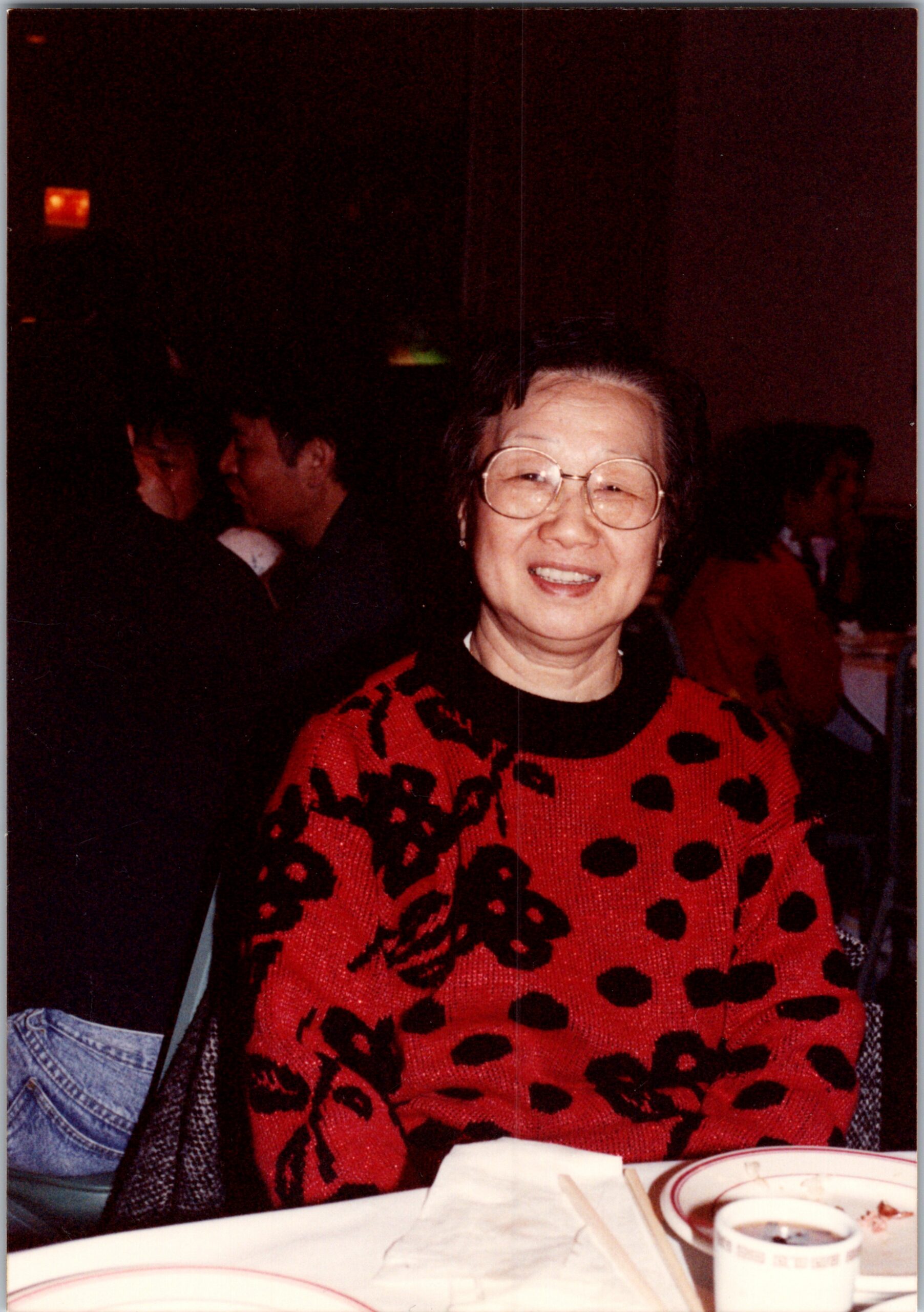 Smiling elderly woman with round glasses wearing a red leopard-print sweater at a restaurant table.