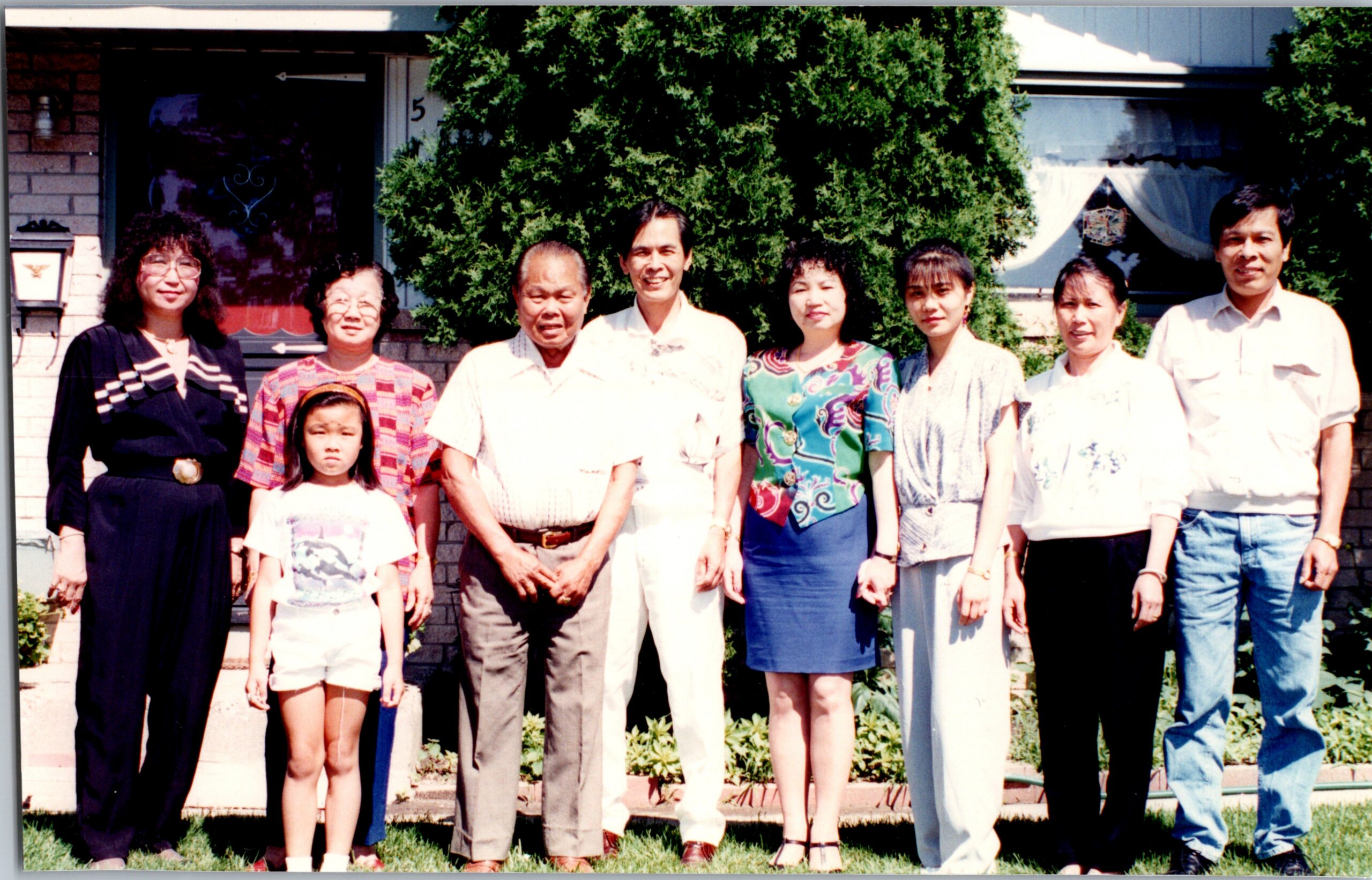 Group photo of a multi-generational family standing in a front yard in front of a house and trees, smiling at the camera.