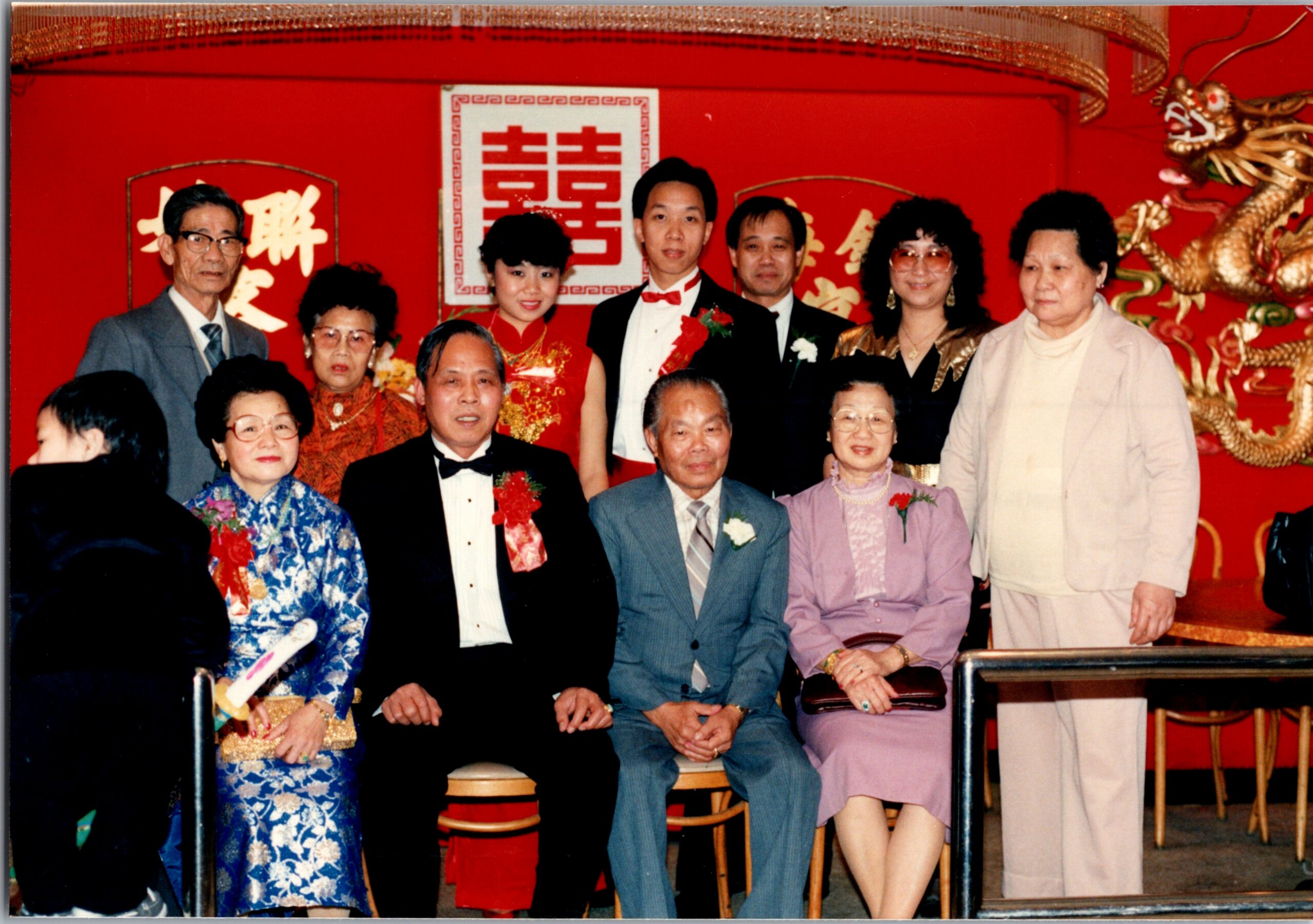 Group of wedding guests in formal attire posing in front of a red Chinese wedding backdrop with a 'Double Happiness' sign and dragon decor.