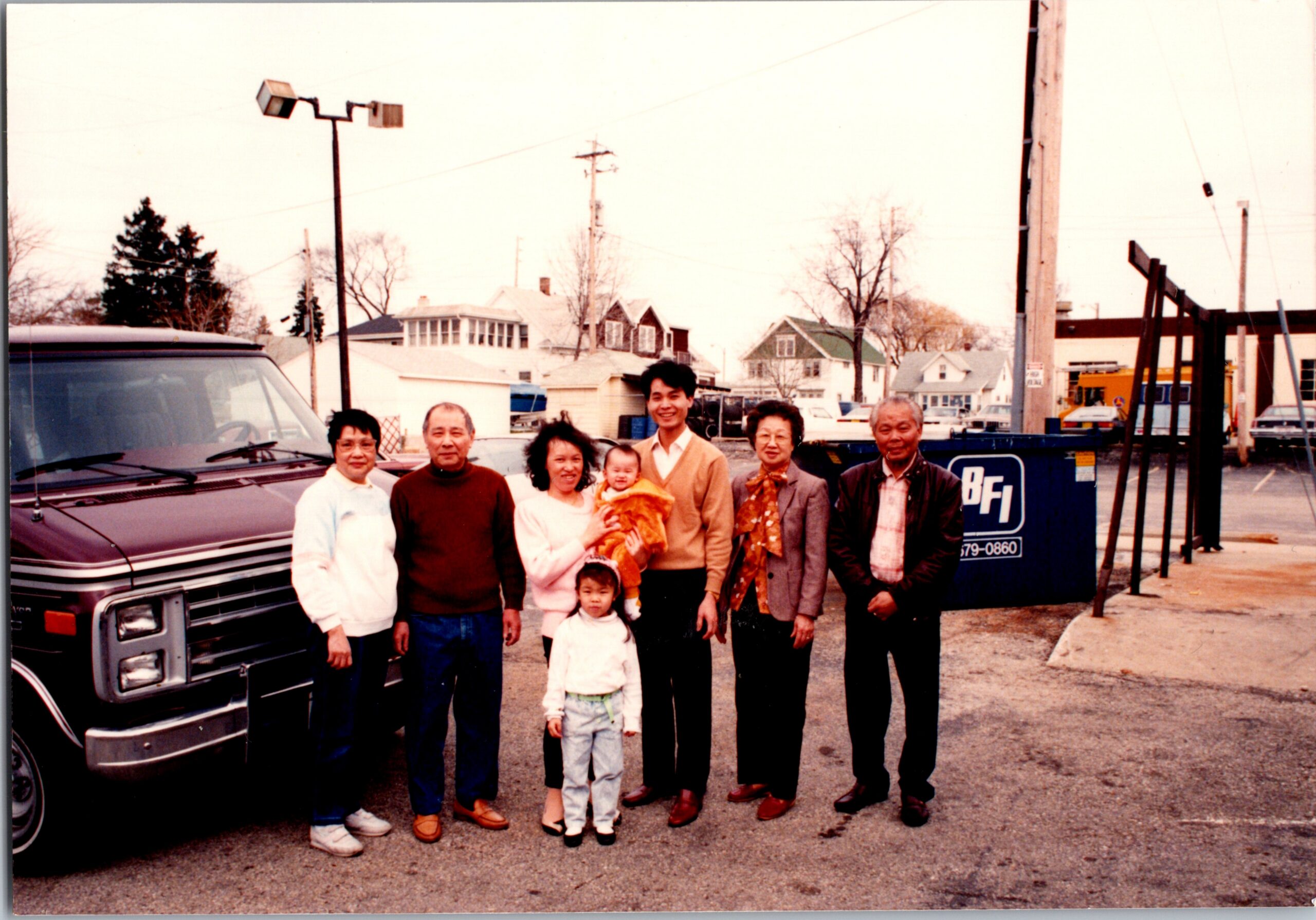 A multi-generational Asian American family posing for a photo in a parking lot beside a large dark van, with houses and a construction site in the background.