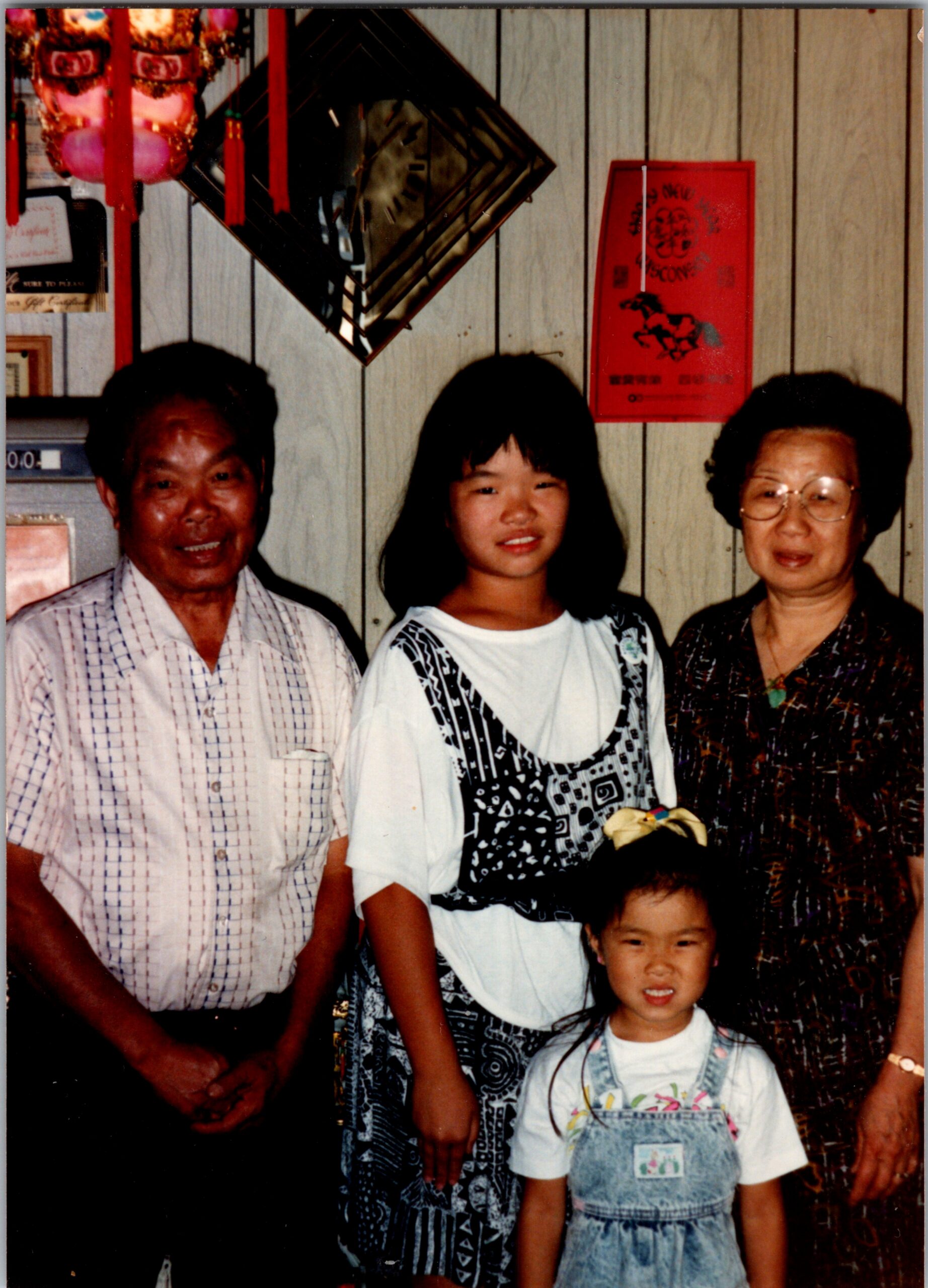 Family group indoors: an older man, a young girl, an older woman, and a child in front, standing against wood-paneled walls.