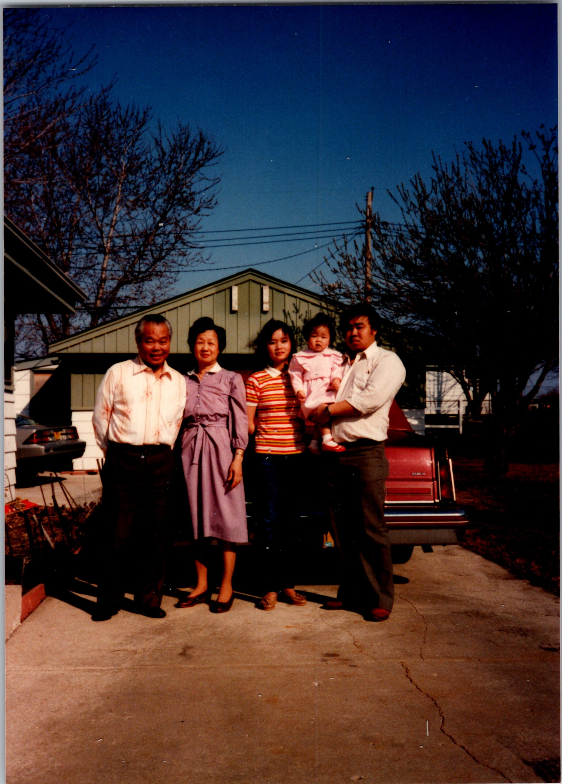Family posing in a driveway in front of a suburban house during daytime, with a baby held by a man on the right.