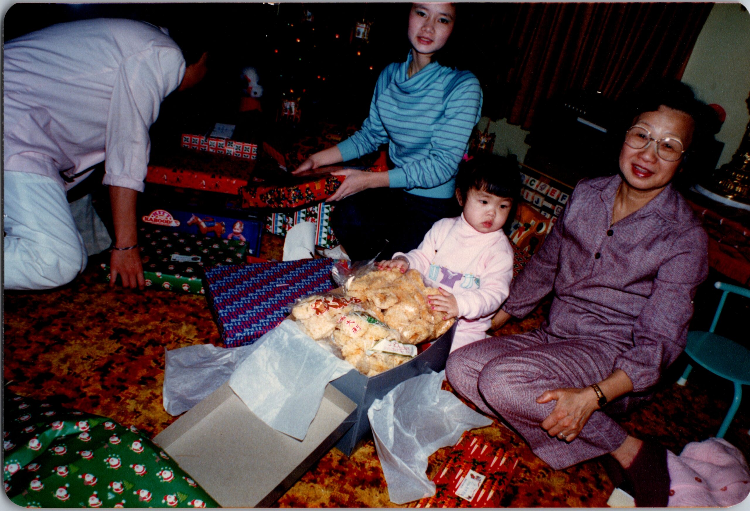 Family members unwrap Christmas gifts around a decorated tree; a toddler sits amid wrapped presents as an adult smiles nearby.