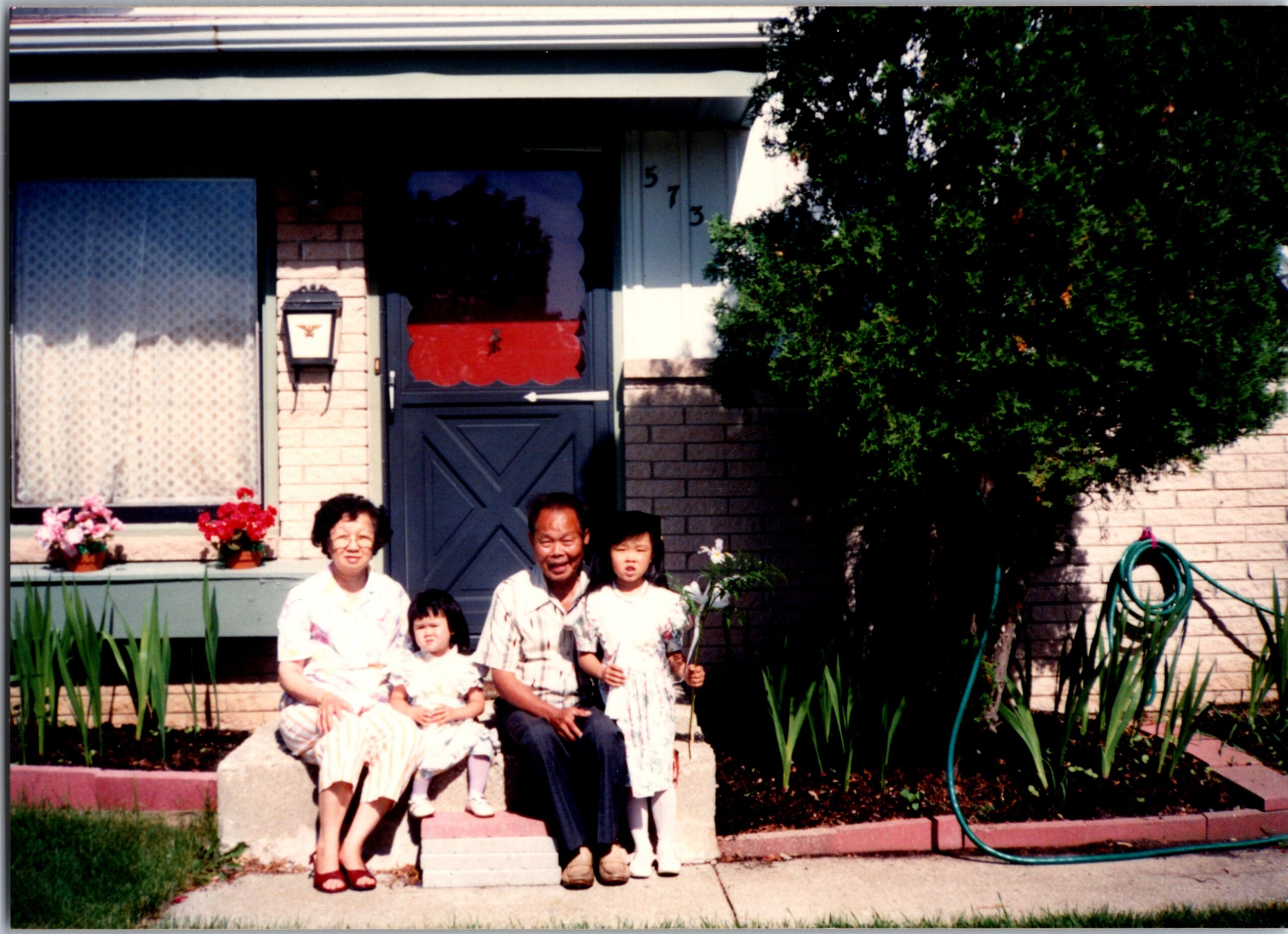 A family of five seated on front steps of a brick house, with a flower bed, lace curtains in the window, and a green hose visible on the right.