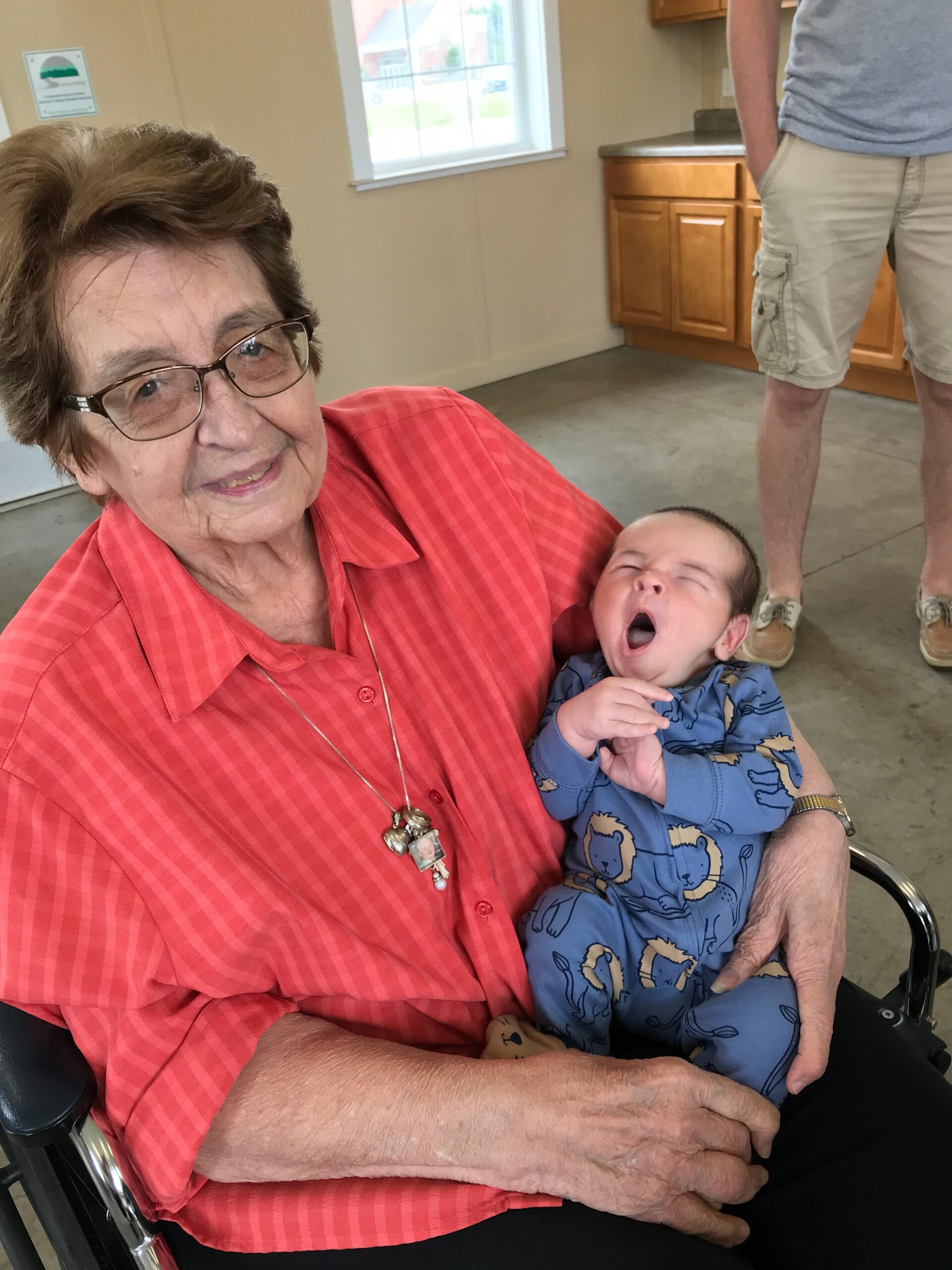 Older woman in a red striped shirt seated in a wheelchair, holding a yawning baby in a blue dinosaur onesie indoors, with a man in shorts visible in the background.