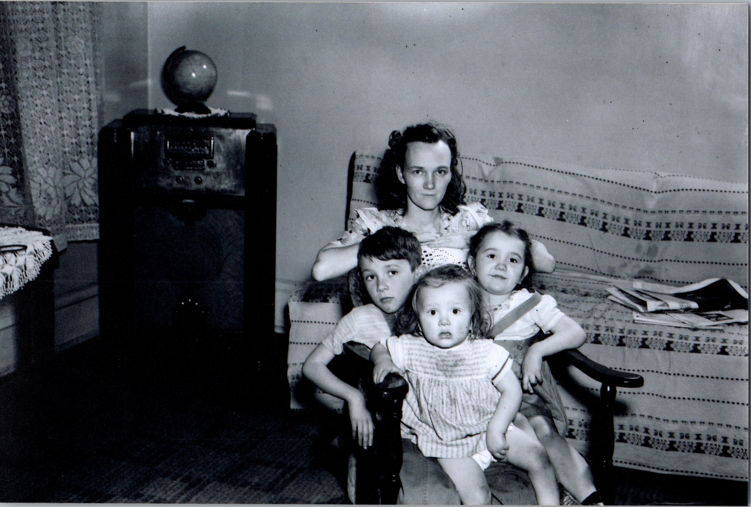 Mother sits with four children on a striped sofa in a vintage living room; globe on a cabinet behind them.