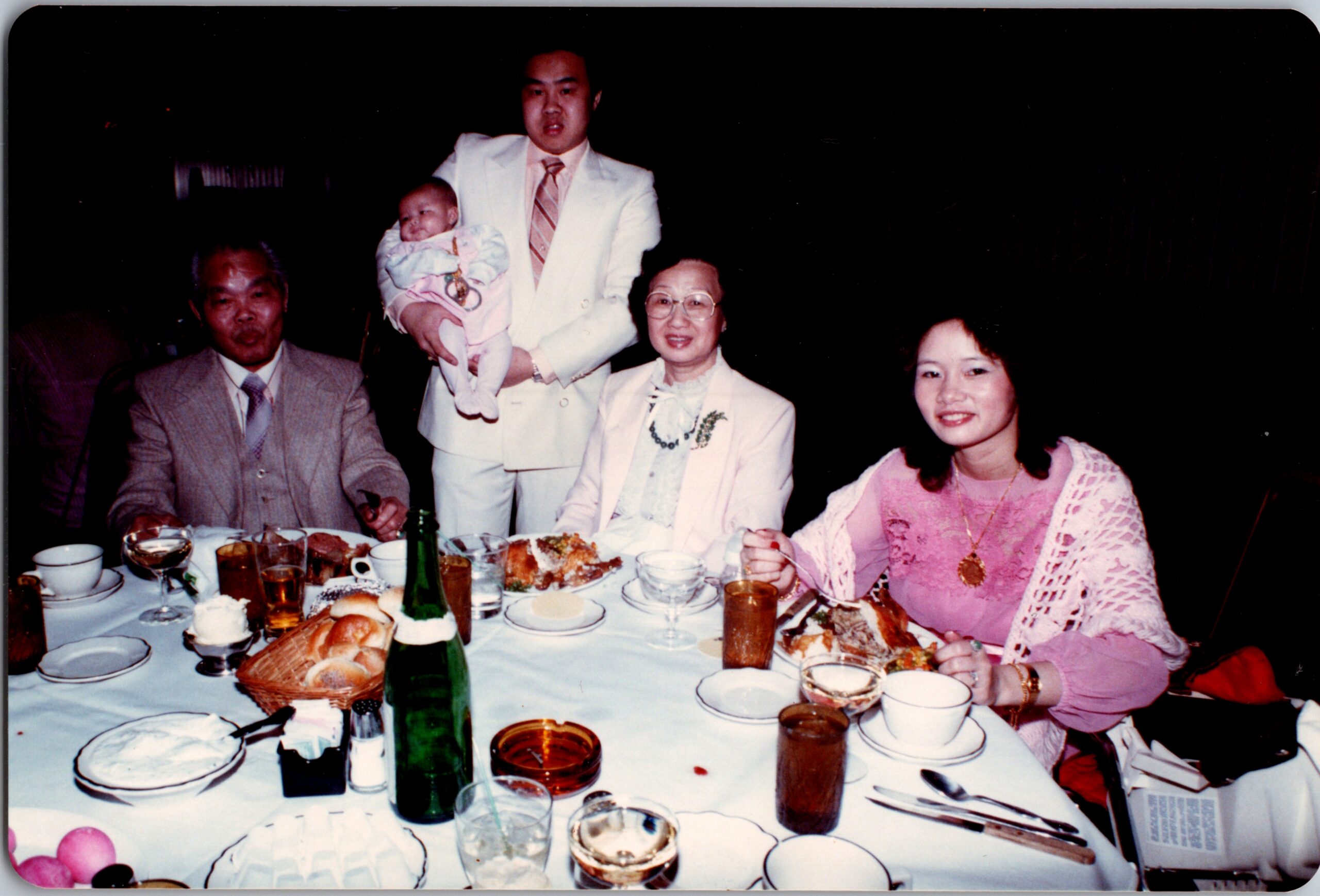 Family gathered at a banquet table; man in a white suit holds a baby while seated adults smile nearby.
